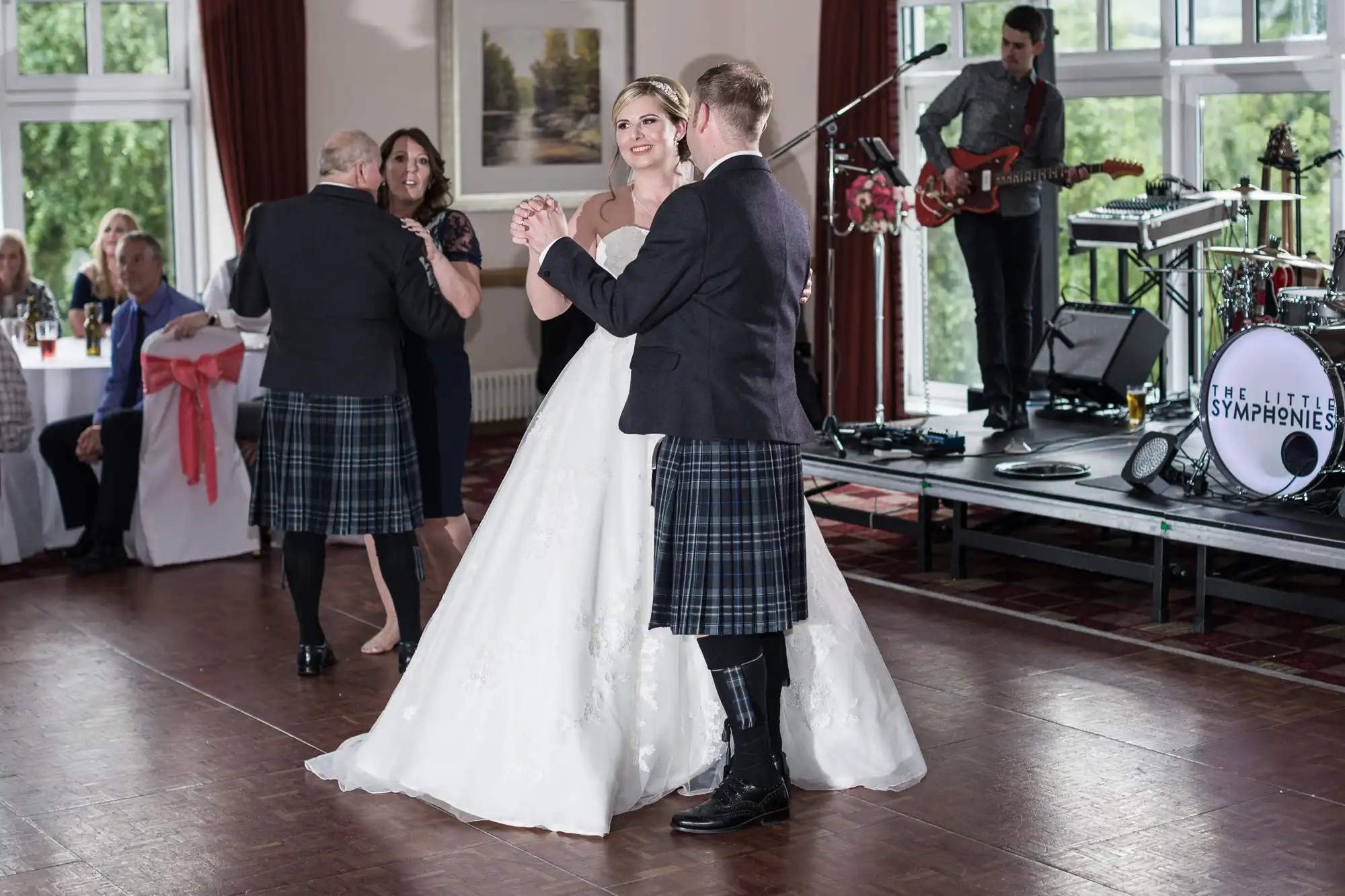 A bride in a white gown dances with a groom in a kilt on a wooden floor. Another couple dances nearby. Guests are seated at tables, and a band with a guitarist and drum kit plays in the background. Large windows let in natural light.