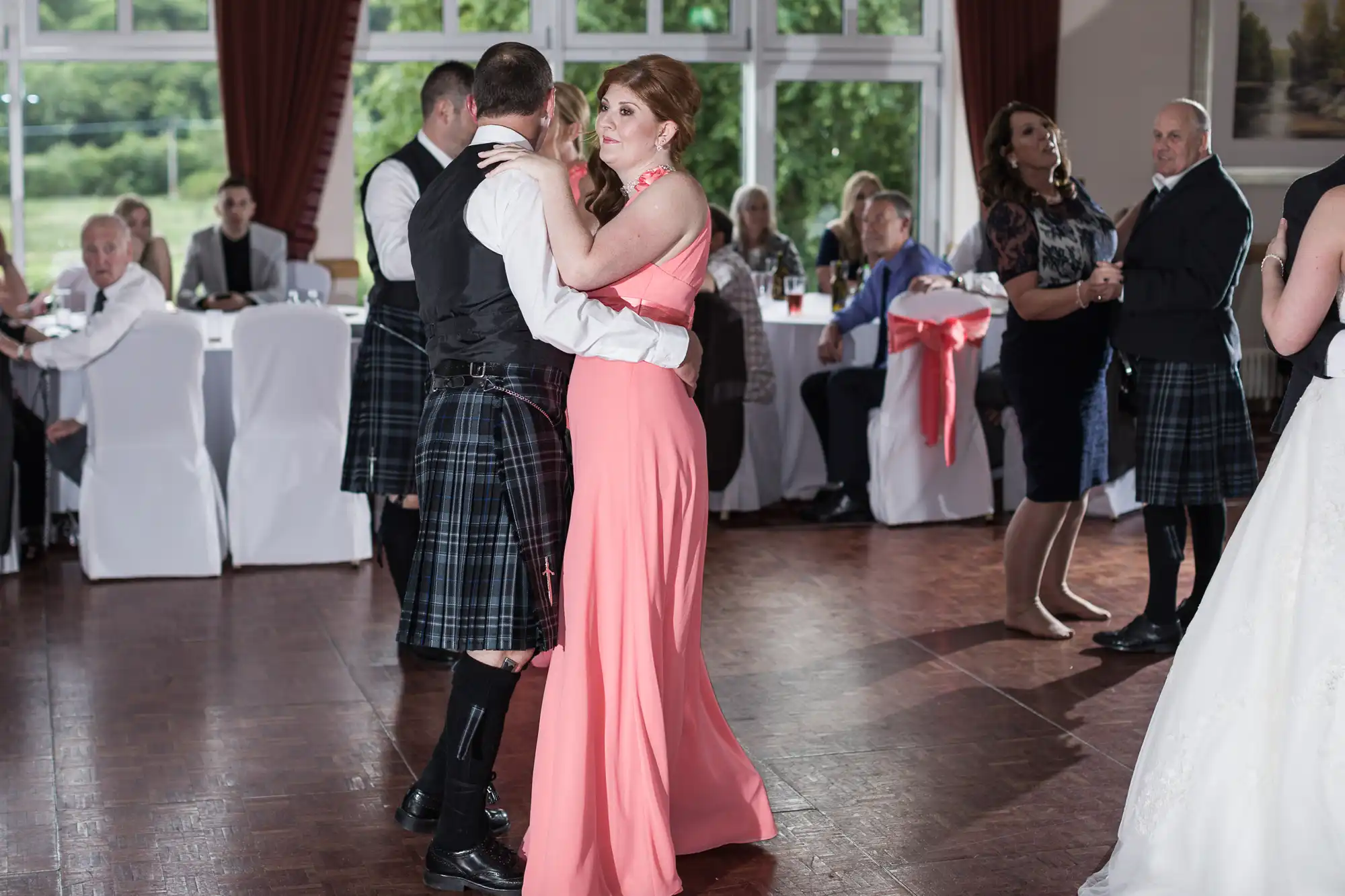A man in a kilt and waistcoat dances with a woman in a long peach dress at a wedding reception. Other guests sit at round tables or dance nearby. Large windows reveal greenery outside, and the room is decorated with white tablecloths and red sashes.