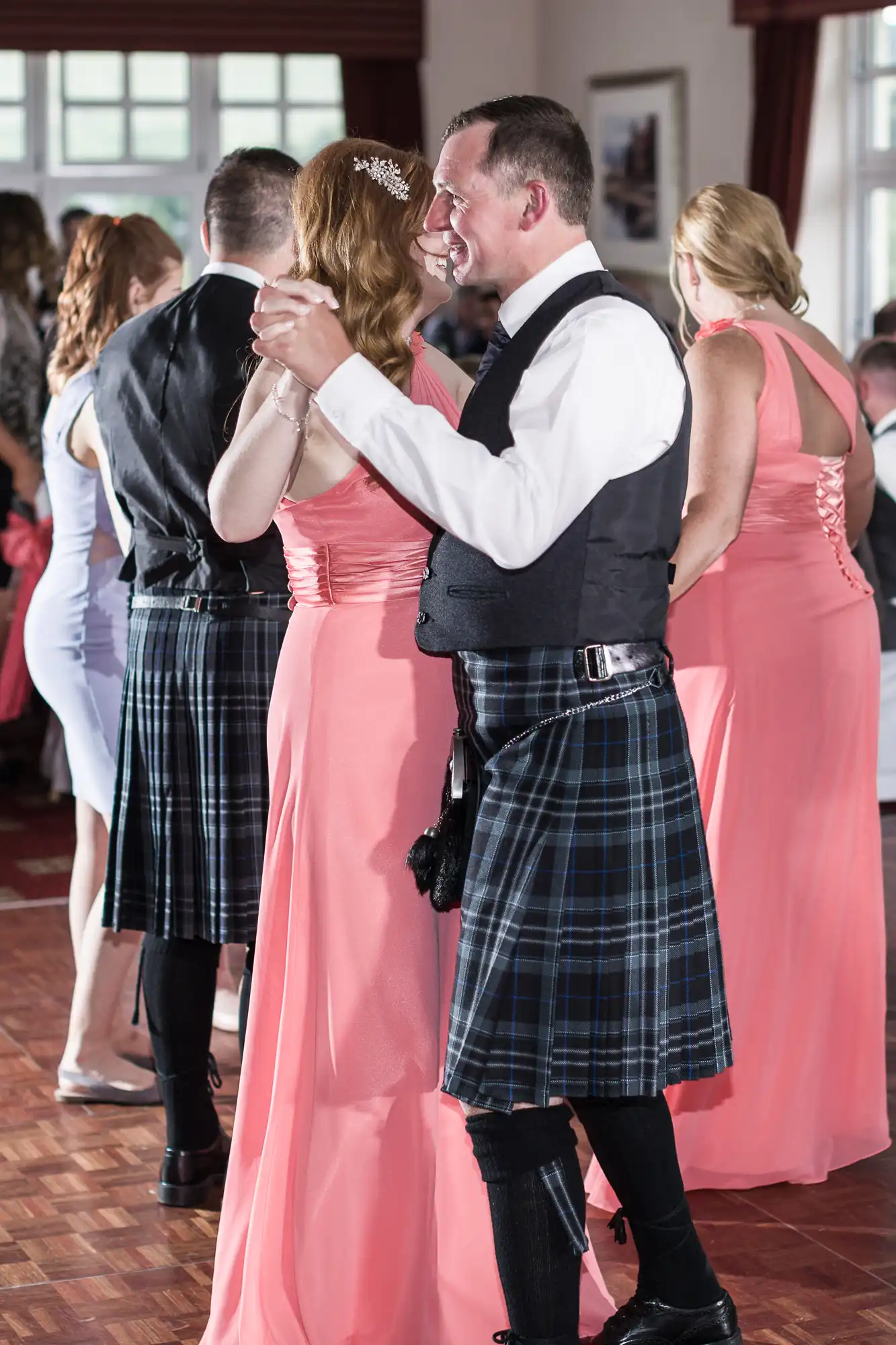 A man in a black waistcoat, white shirt, and tartan kilt dances and smiles with a woman in a long, pink dress. Other guests in formal attire, including kilts and pink dresses, dance in the background at a bright indoor event.