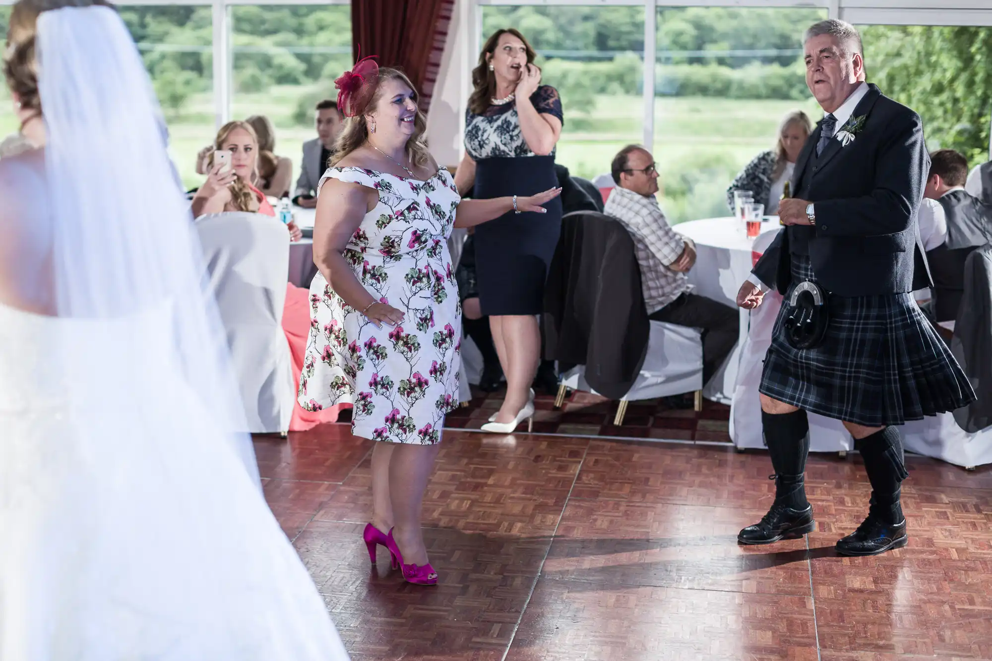 A woman in a floral dress and pink shoes dances with a man in a kilt on a wooden floor at a wedding reception. Guests sit at round tables in the background, some watching, with windows revealing a green landscape outside.