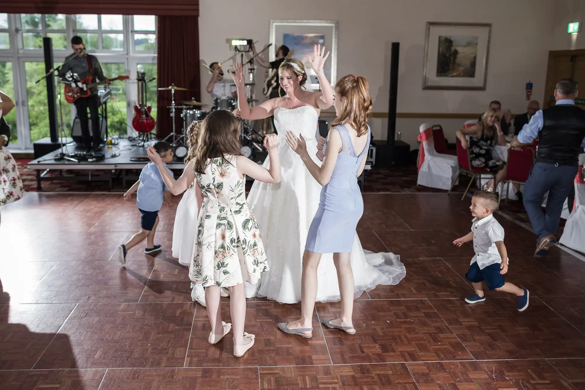 A bride in a white dress dances joyfully with children and a woman in a light blue dress on a wooden dance floor at a wedding reception, whilst a live band plays on stage in the background. Guests are seated at tables, watching and smiling.