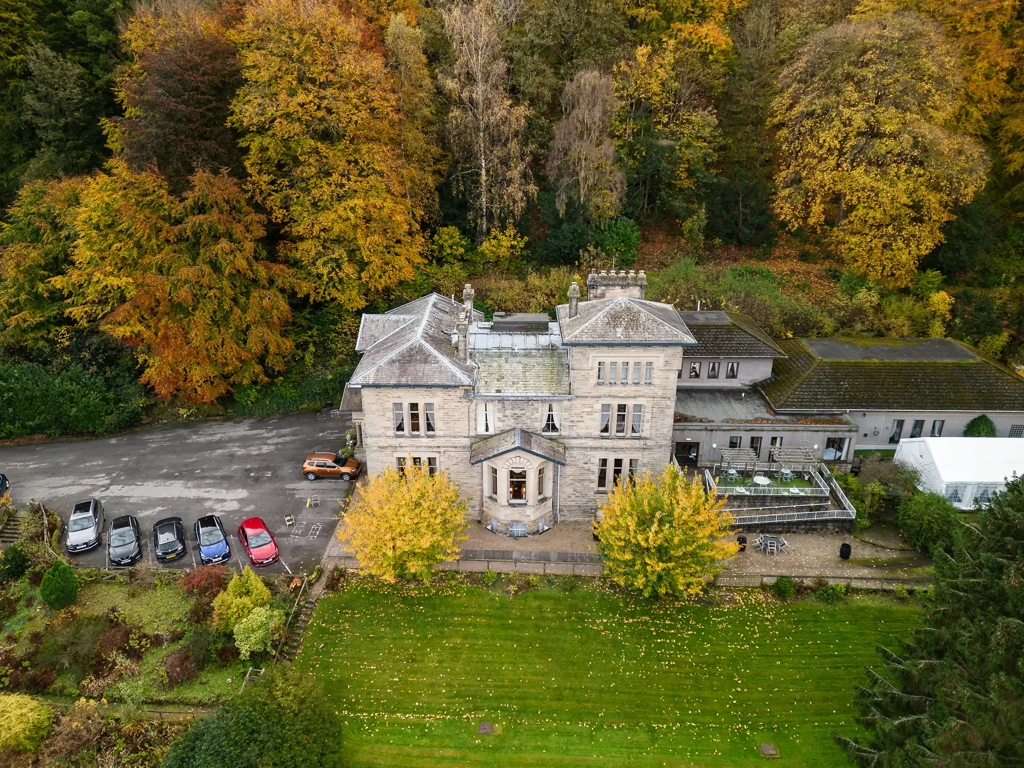 A large stone manor house with a slate roof sits amid autumn trees. Six cars are parked on a car park beside it. The manicured lawn in front is scattered with yellow leaves, and the wooded hillside behind is filled with vibrant autumn foliage.