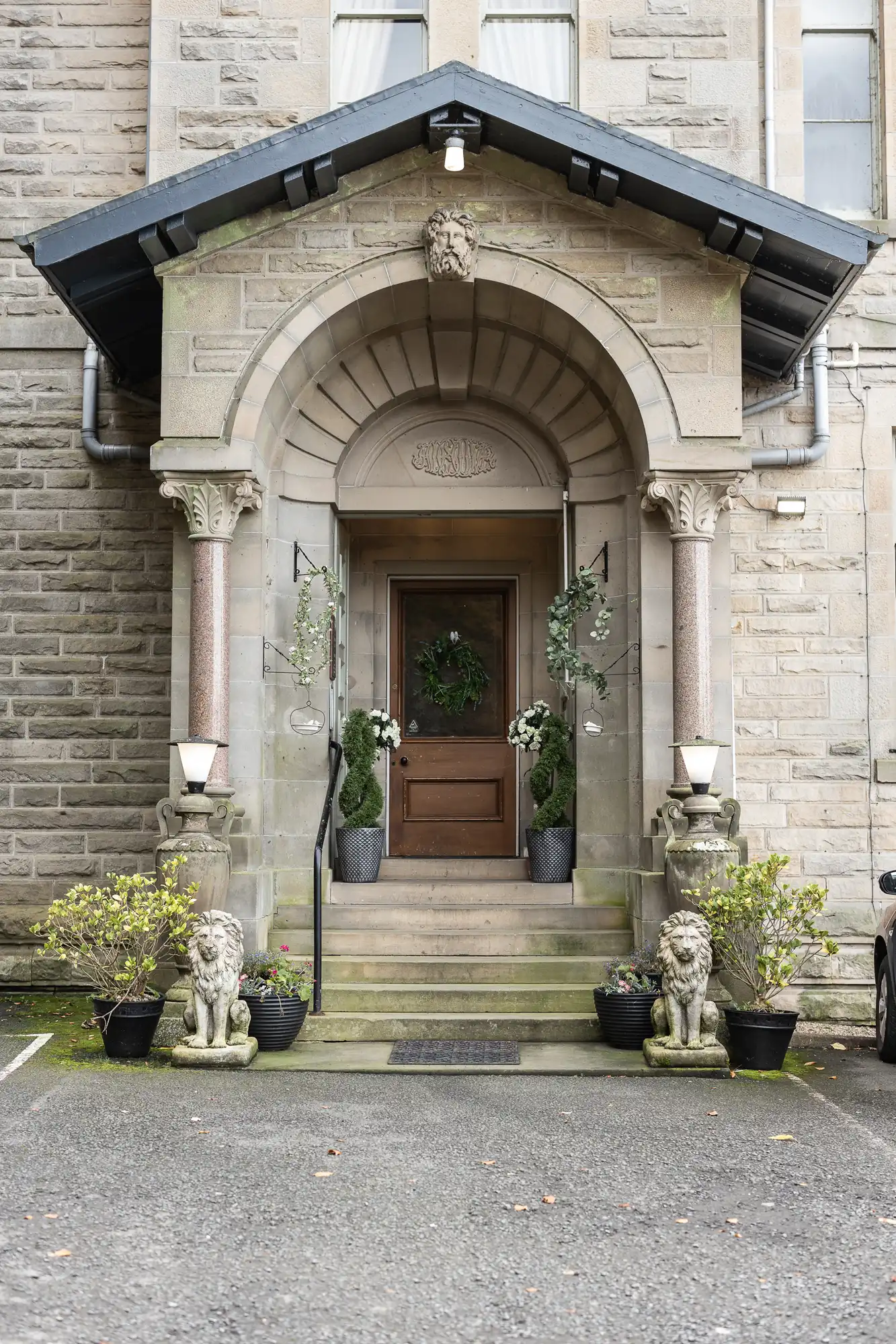 A stone building entrance with an arched doorway, two columns, a wooden door, and potted plants. Two stone lion statues sit at the base of steps leading up. Hanging planters and greenery flank the entrance; the building exterior is textured stone.