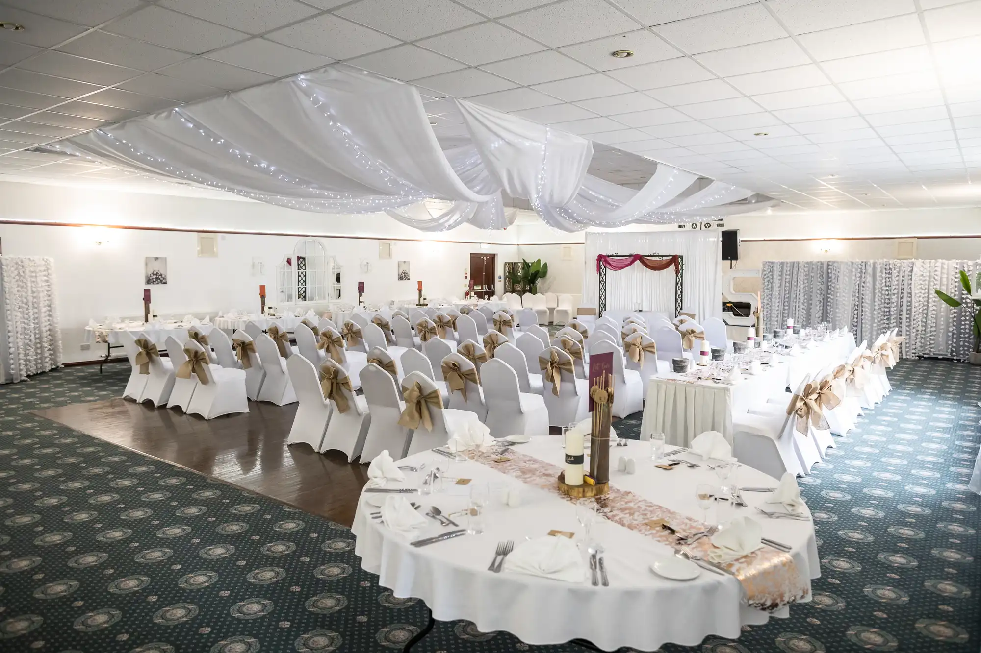 A spacious banqueting hall decorated for an event, with white-clothed tables and chairs draped in white covers and beige sashes. Ceiling features flowing white fabric and fairy lights. Tables are set with plates, cutlery, and centrepieces. The floor is carpeted.