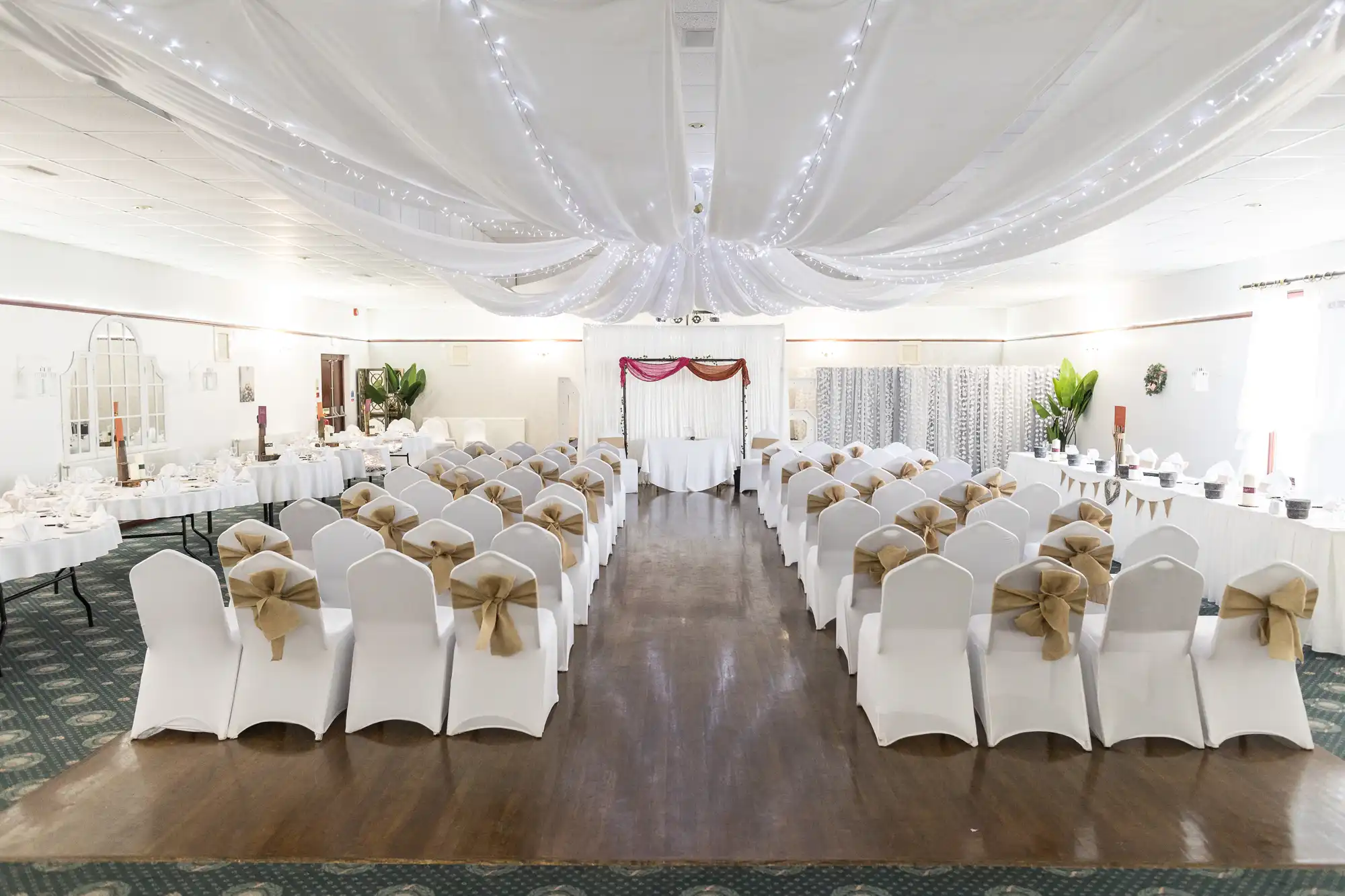 A banqueting hall set for a wedding, with rows of white chairs adorned with beige bows facing an altar draped in white and red fabric. Ceiling is decorated with white drapes and fairy lights; tables are set along the sides, and plants decorate the room.
