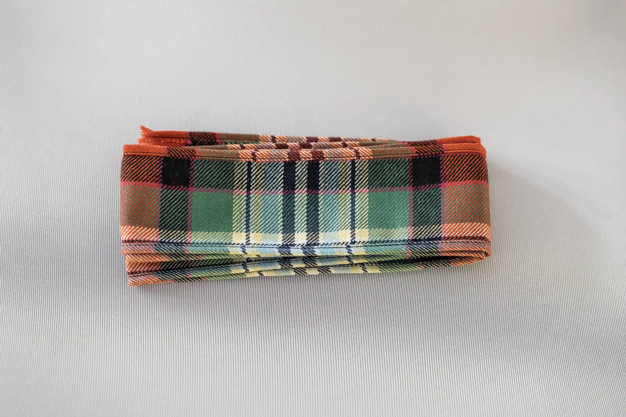 A folded fabric item with a tartan check pattern featuring green, orange, yellow, and black stripes, placed on a light grey background. The fabric is neatly stacked, with visible crisp edges and evenly aligned folds.