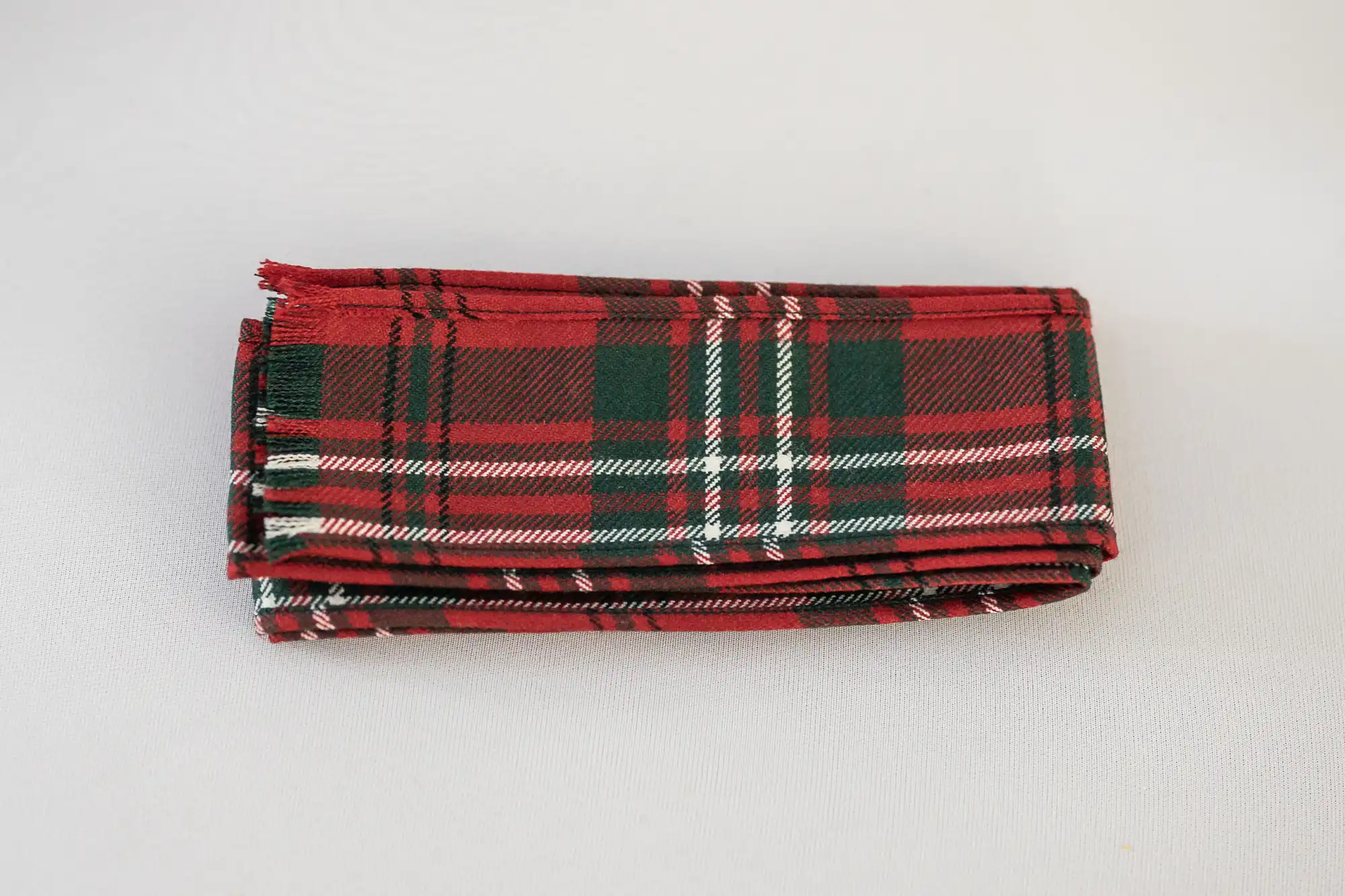 A neatly folded, rectangular piece of fabric with a traditional red, green, white, and black checked tartan pattern, displayed on a plain white surface. The fabric edges are slightly frayed, giving it a textured look.