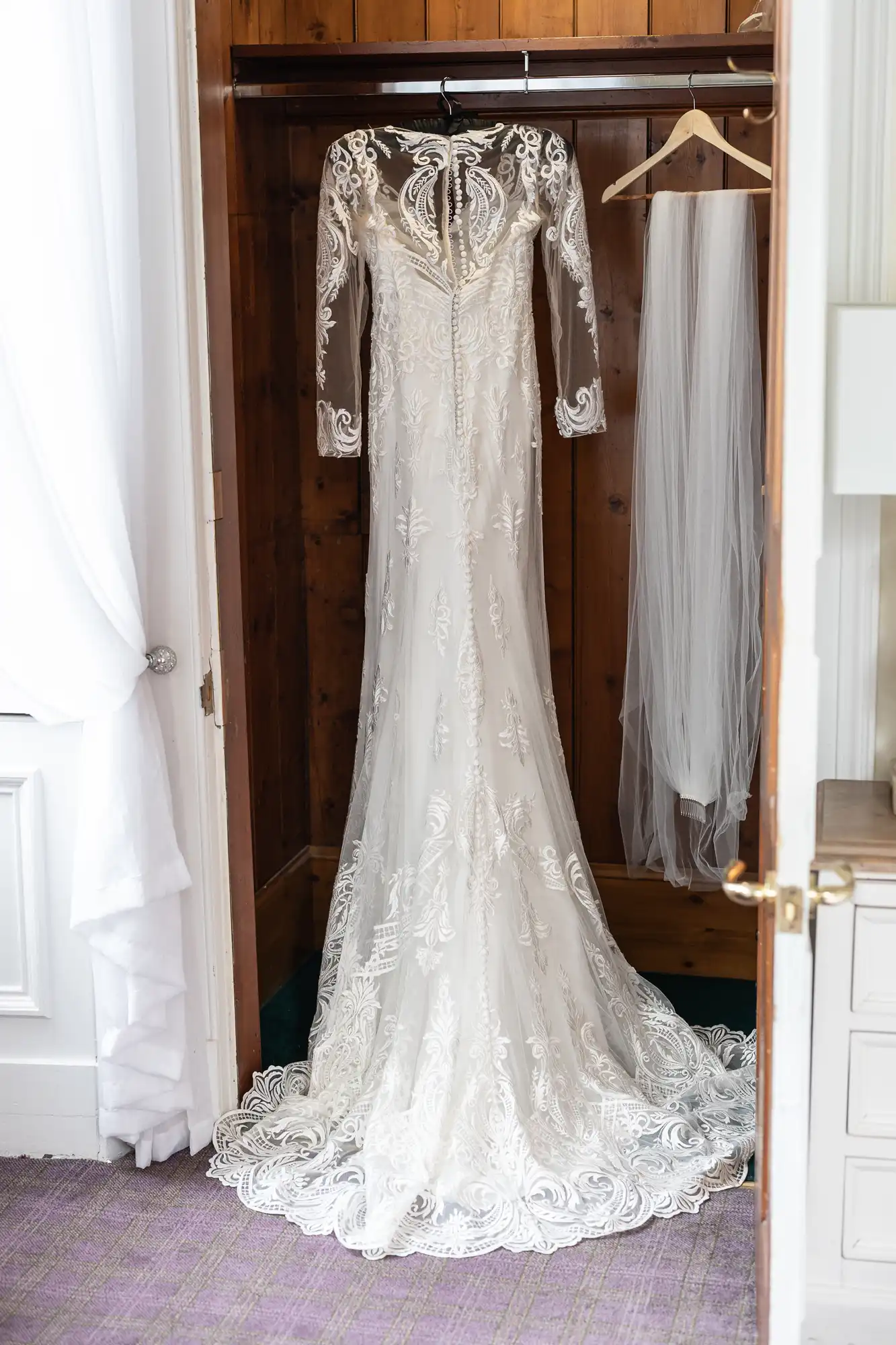 An elegant, long-sleeved lace wedding dress with a train hangs inside a wooden wardrobe. The gown features intricate embroidery and sheer sections. Beside it, a white veil hangs on a hanger. The wardrobe stands open in a bright, softly lit room.