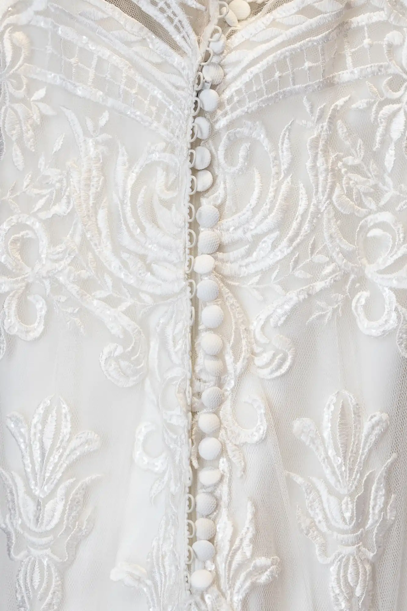 Close-up of a white lace fabric featuring ornate floral embroidery, with a row of small white fabric-covered buttons running vertically down the centre. The texture and detail of the lace and buttons are clearly visible.
