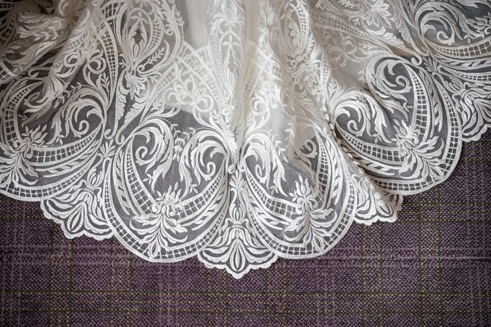 A close-up of an ornate white lace fabric with intricate floral and scroll patterns, scalloped edges, and delicate embroidery, draped over a purple checked textured surface.