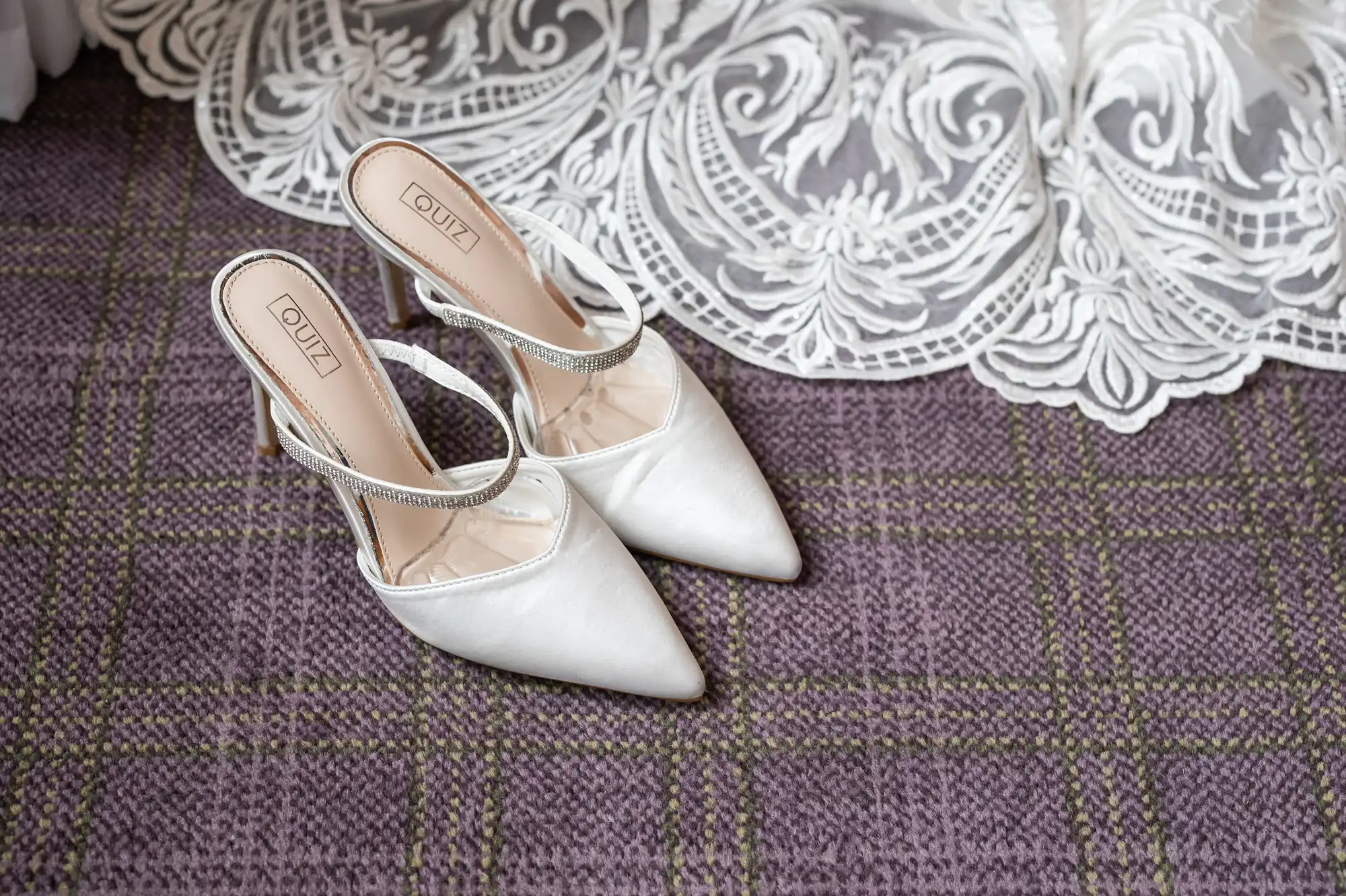 A pair of elegant white pointed-toe heeled shoes with a rhinestone strap sits on a purple checked carpet next to a lacy white bridal dress hem. The shoes are branded "QUIZ" and placed neatly side by side.