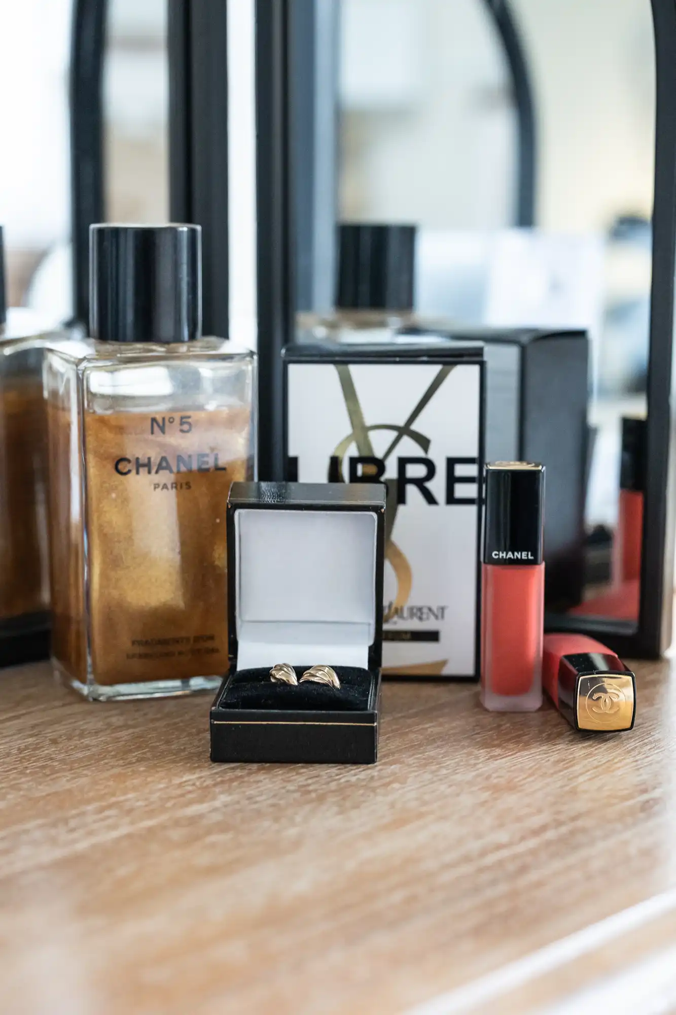 A bottle of Chanel No. 5 perfume, Yves Saint Laurent Libre perfume, a black lipstick tube, a red Chanel lip gloss, and an open jewellery box with gold earrings are arranged on a wooden surface in front of a mirror.