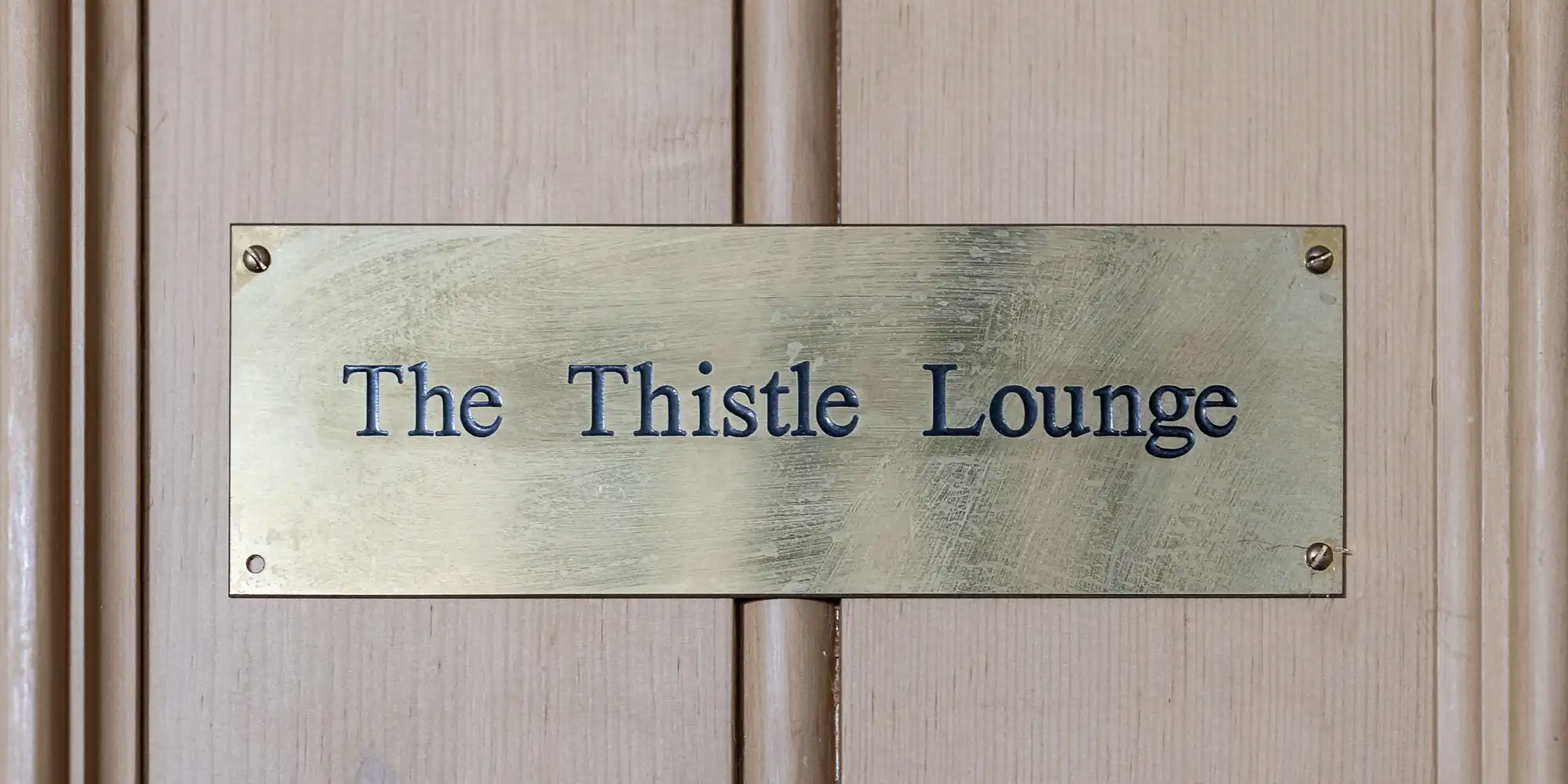 A brass plaque with engraved black text reading "The Thistle Lounge" is mounted on a light wooden door. The plaque is rectangular with bevelled edges and secured by two visible screws, one on each side.