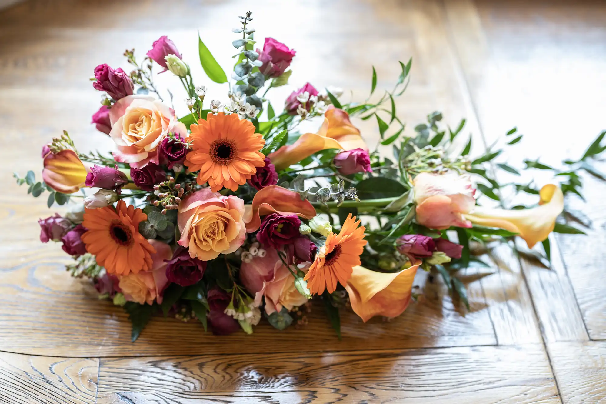 A colourful floral bouquet rests on a wooden surface, featuring orange gerbera daisies, yellow calla lilies, peach roses, purple bi-coloured roses, eucalyptus leaves, and green foliage, arranged in a natural, slightly cascading style.