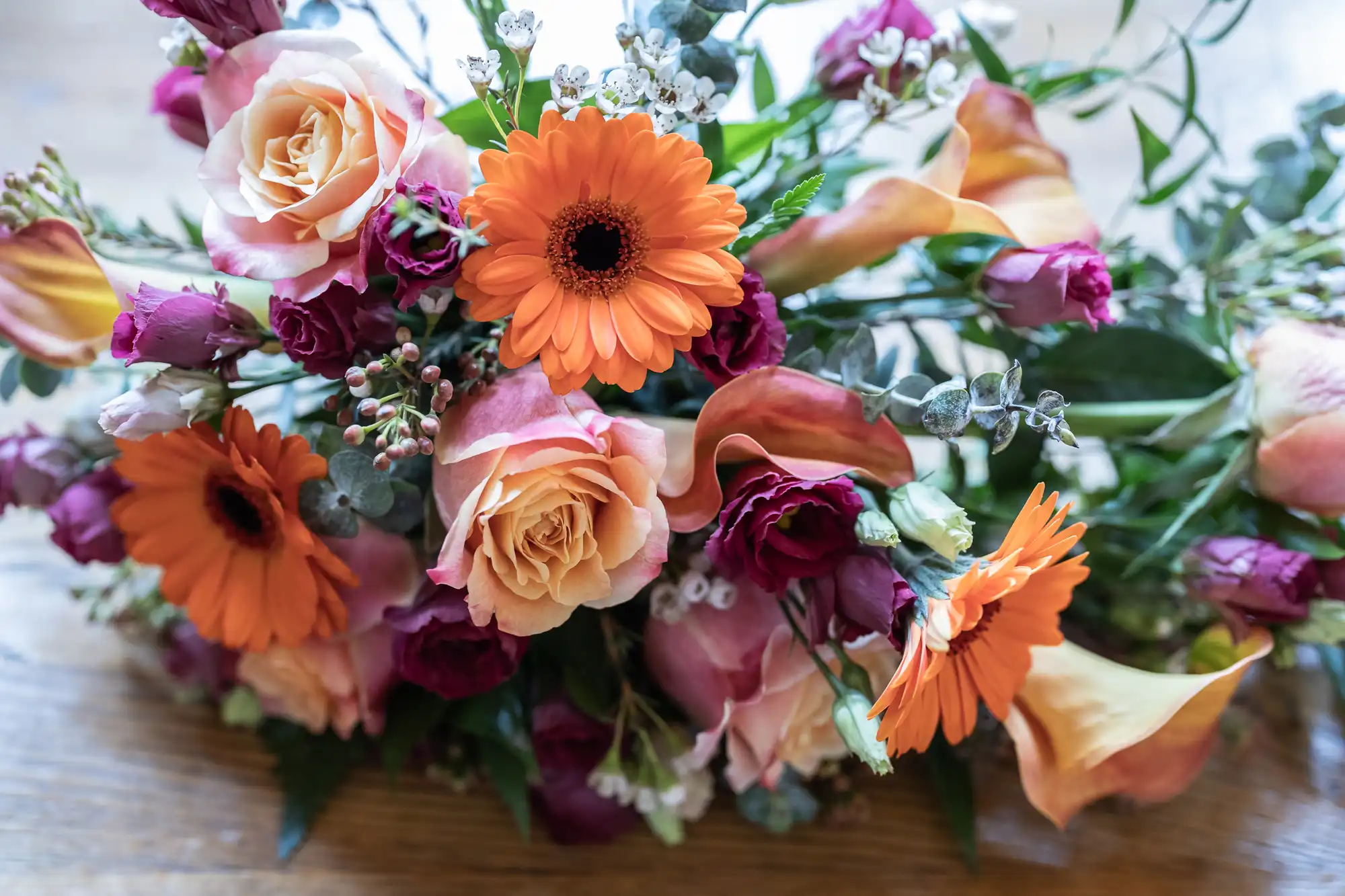 A vibrant bouquet with orange gerbera daisies, peach and pink roses, deep magenta roses, calla lilies, sprigs of eucalyptus, and small white flowers, arranged on a wooden surface. The flowers are fresh and lush, with varied textures and colours.