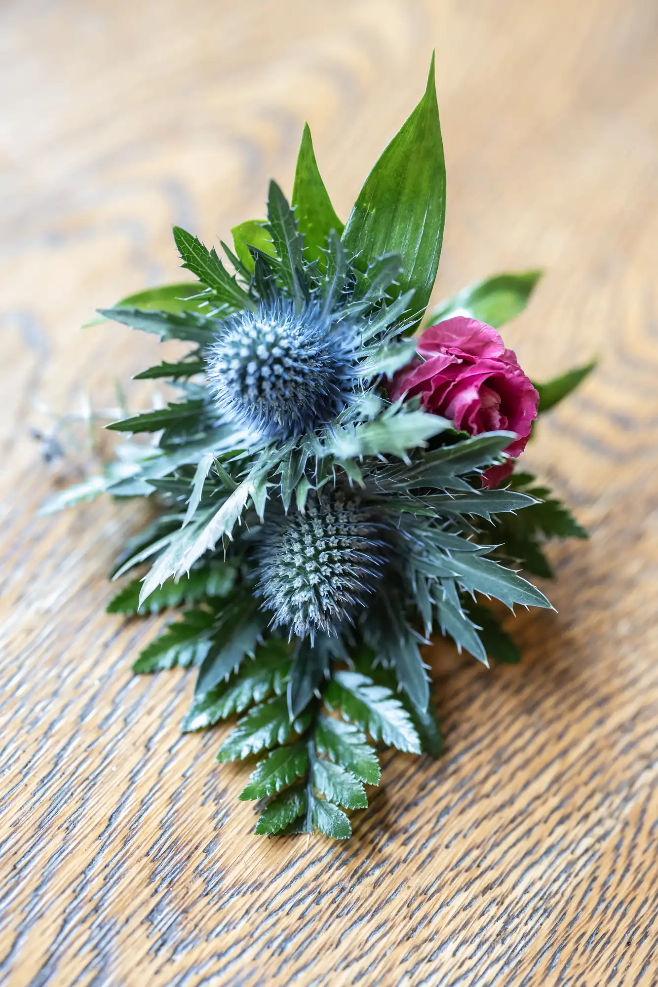 A small floral arrangement with two spiky blue thistles, a single deep pink rose, and green foliage, resting on a light brown wooden surface with a visible grain pattern.