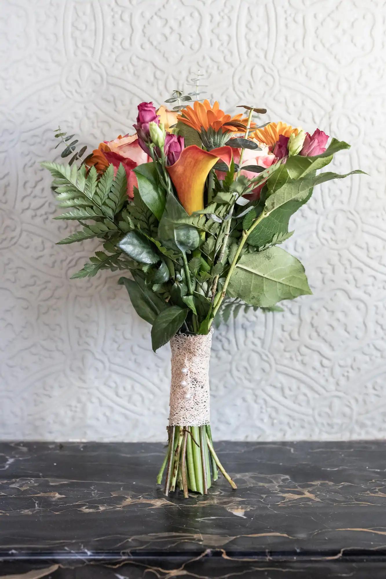 A bouquet of assorted flowers with pink and orange blooms, calla lilies, and greenery, tied with a band of lace, stands upright on a dark stone surface against a light grey, textured, patterned wall.