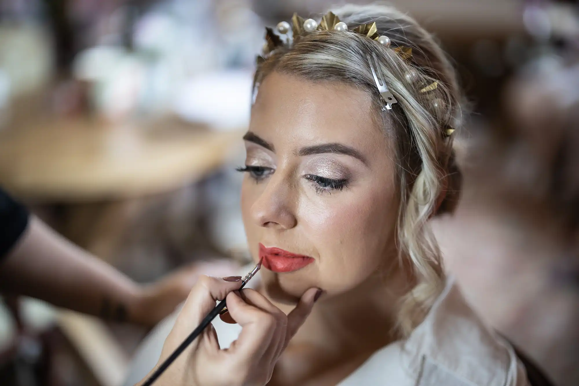 A woman with blonde hair styled with a gold and pearl hairband is having red lipstick applied by a make-up artist. She wears a white top and has soft, natural make-up with shimmer on her eyelids. The background is blurred, focussing on her face.