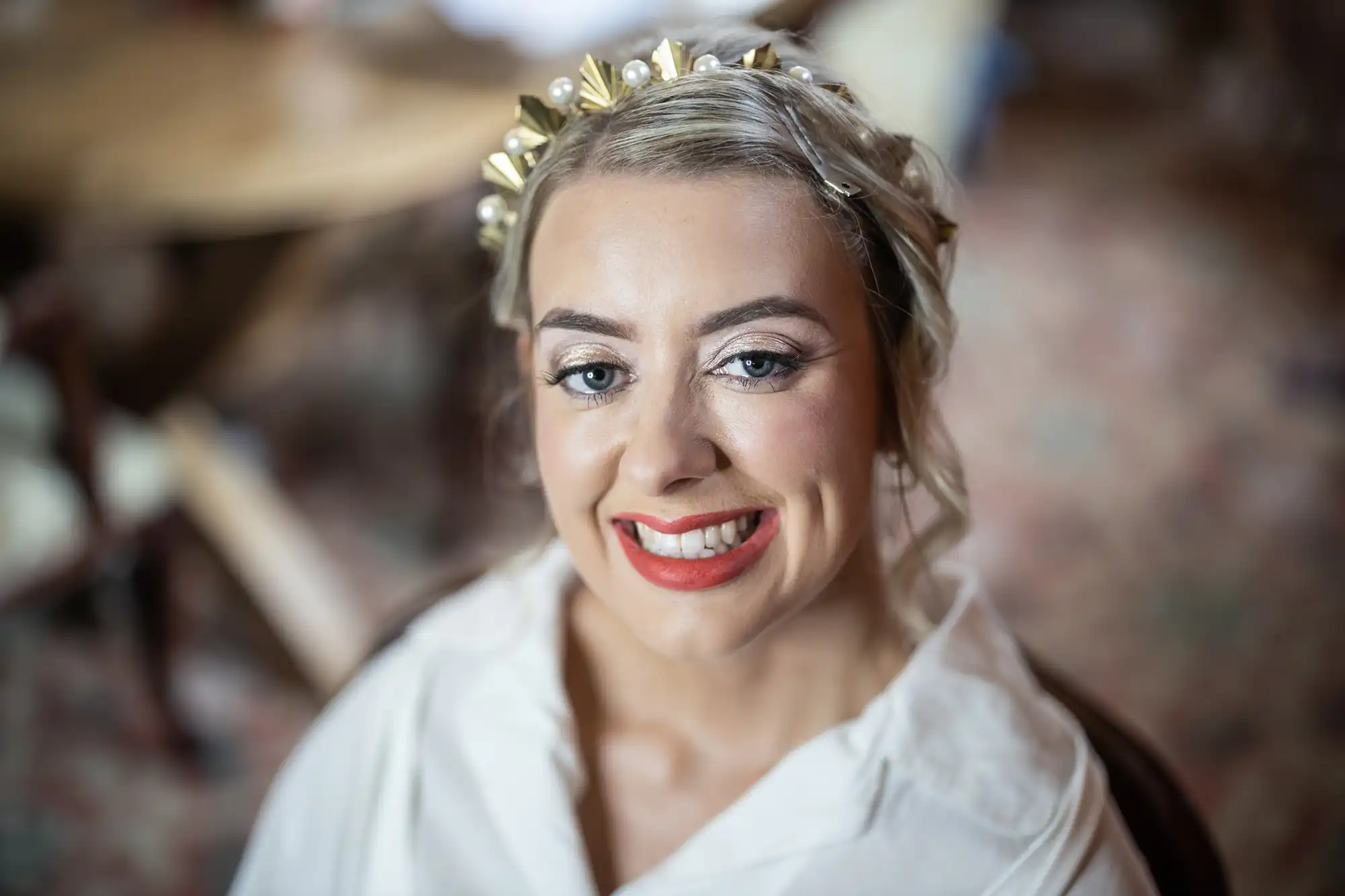 A woman with blonde hair styled in an updo and adorned with a gold accessory smiles brightly. She wears bold red lipstick, eye make-up, and a white outfit. The background is blurred, showing indoor wooden furniture and a patterned carpet.