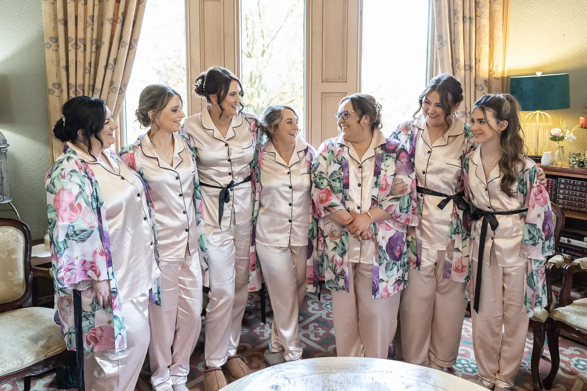 Seven women stand in a row indoors, all wearing matching light pink satin pyjamas with black trim; six wear floral dressing gowns. They smile and face each other, appearing happy and relaxed, suggesting a pre-wedding or celebratory gathering.