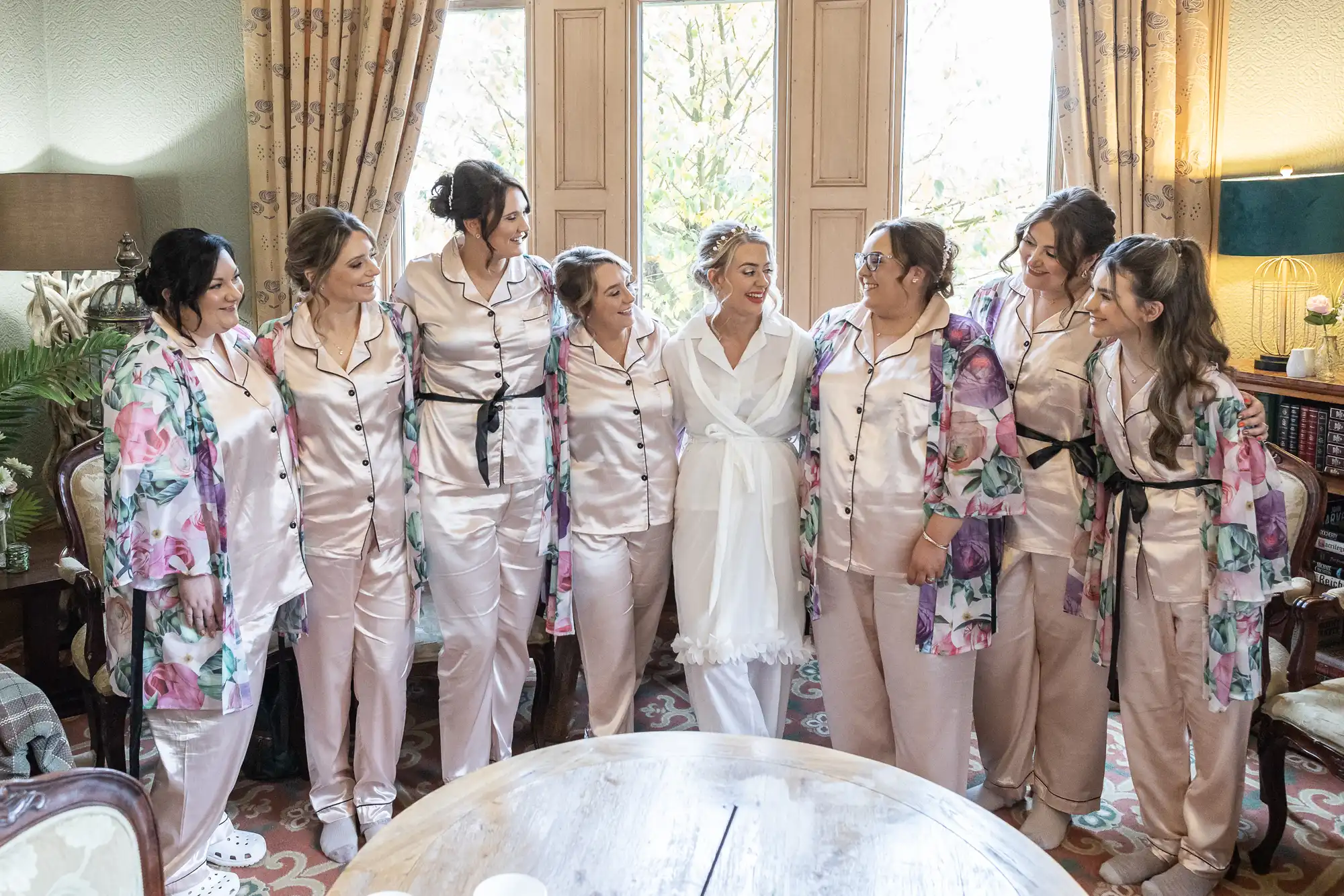 Eight women stand smiling together indoors, all wearing light pink satin pyjamas; the central woman wears a white dressing gown. Some women have floral dressing gowns over their pyjamas. Curtains frame a tall window behind them, and the room has carpet and vintage furniture.