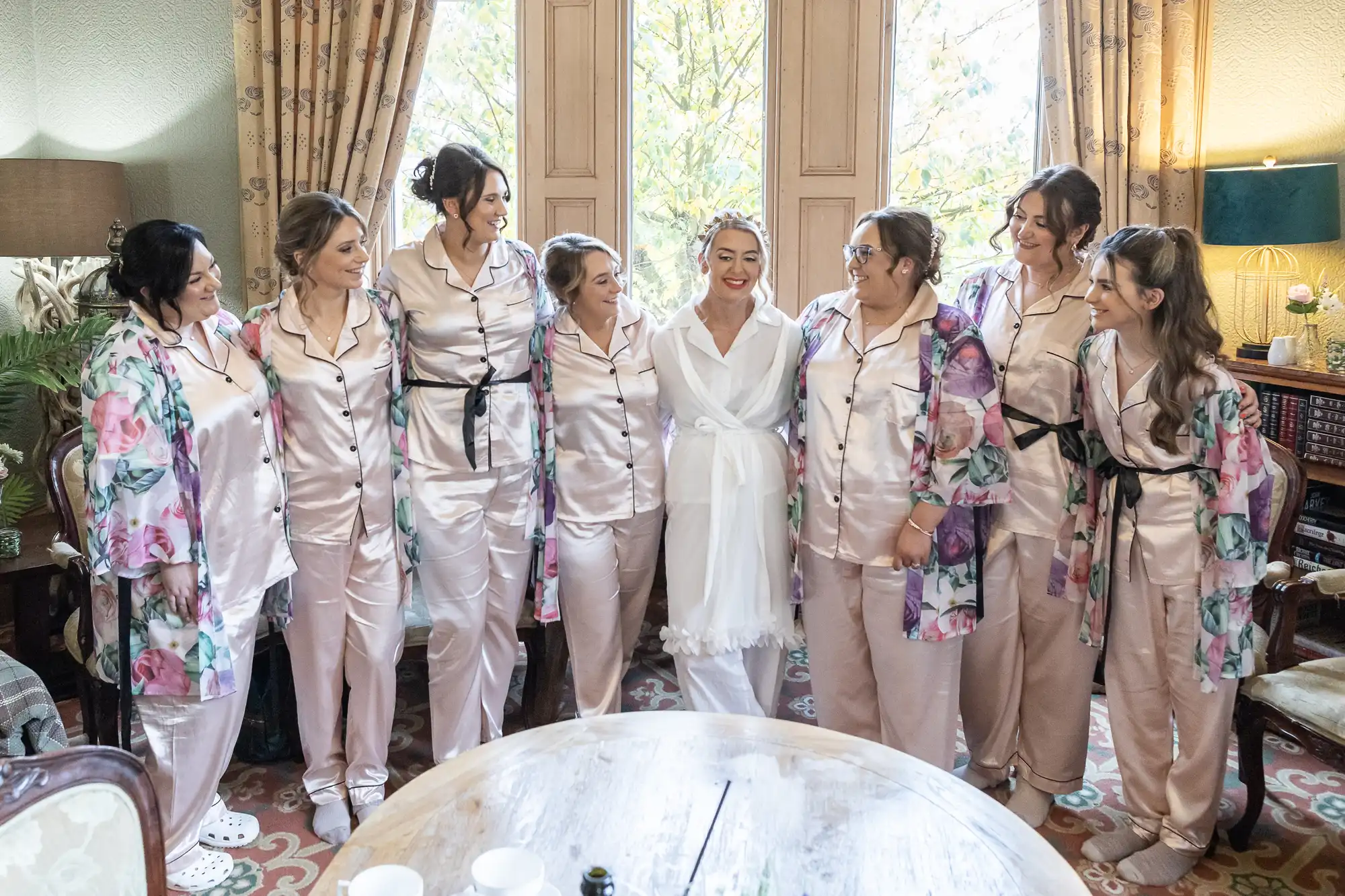 Seven women in matching light pink pyjamas, some with floral dressing gowns, stand smiling in a semicircle around a bride in a white dressing gown. They pose together inside a warmly lit room with large windows and patterned carpet, suggesting a wedding preparation scene.