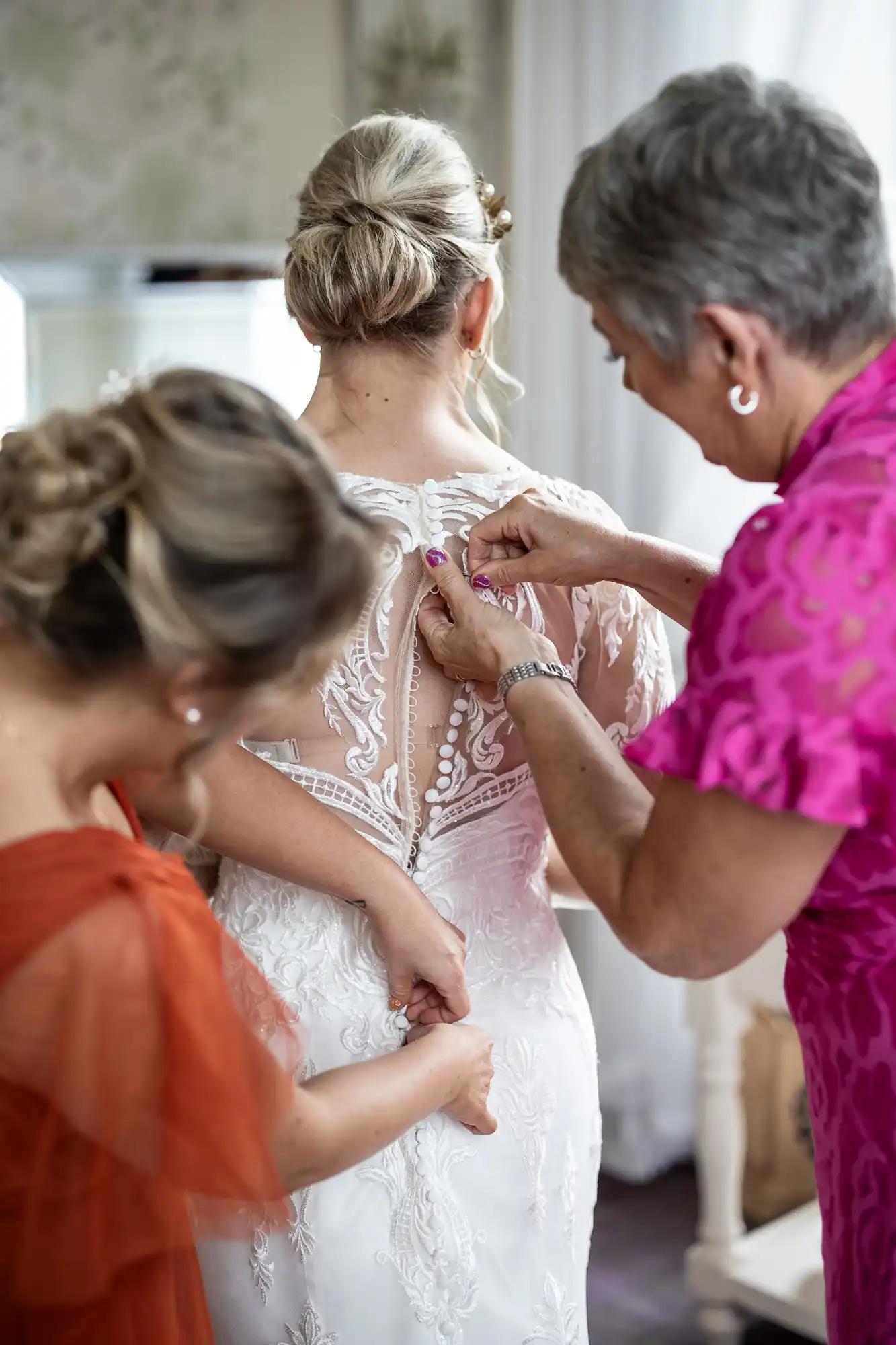 A bride in a white lace wedding dress stands with her back to the camera as two women help to do up her gown. One woman wears a bright pink dress, the other an orange dress. The focus is on their hands and the dress’s detailed lace back.