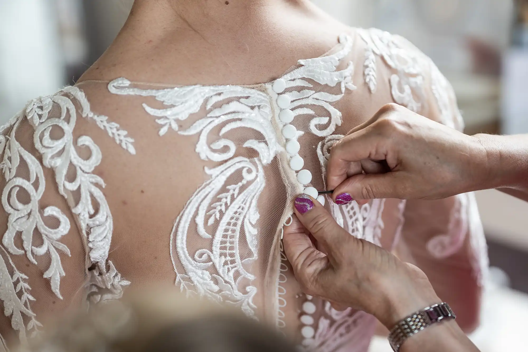 Close-up of hands buttoning the back of a bride’s lace wedding dress with sheer fabric and intricate white embroidery, highlighting the delicate buttons and elegant patterns. The bride’s back and part of her shoulder are visible.