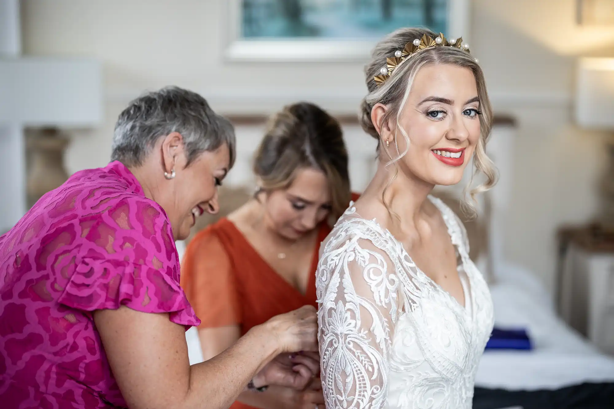 A smiling bride in a lacy white gown stands as two women help fasten her dress. One woman wears a bright pink blouse, the other an orange dress. The bride has blonde hair in an up-do with a gold headband. The background is softly blurred.