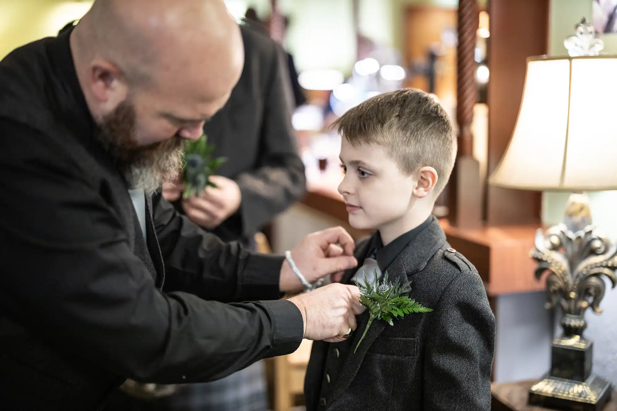 A bearded man pins a buttonhole onto a young boy's dark suit jacket. The boy looks ahead calmly while standing indoors near a lamp and wooden furniture. Both are dressed formally, suggesting a special event like a wedding.