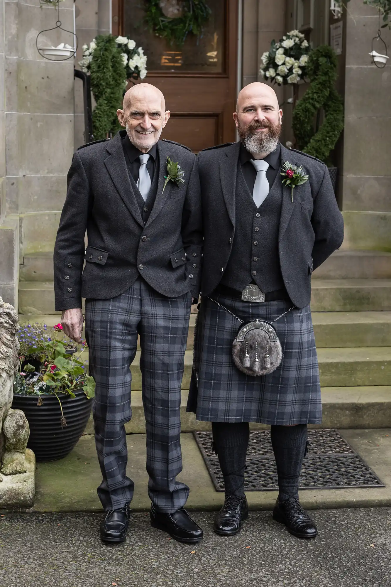 Two men stand side by side in front of stone steps, both wearing formal Scottish attire. The man on the left wears tartan trousers, the man on the right wears a matching tartan kilt with a sporran. Both wear dark jackets, ties, and buttonholes, smiling at the camera.