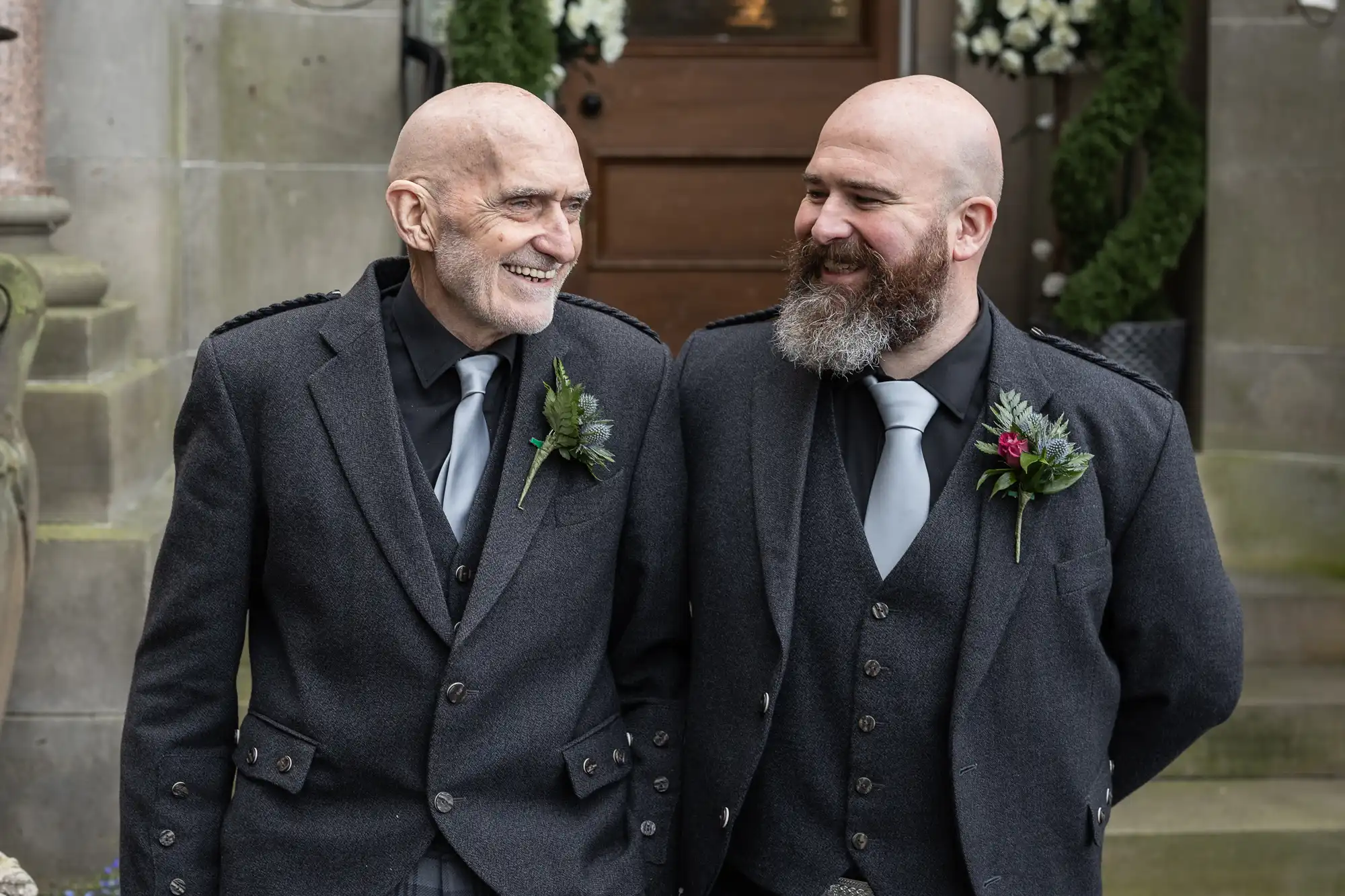 Two bald men with beards, dressed in matching dark suits, grey shirts, and light grey ties, stand close together smiling. Each has a buttonhole on his lapel. They are outside, in front of a stone building with steps and greenery.
