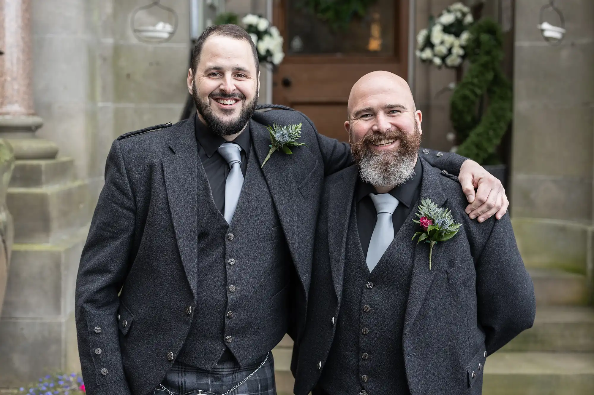 Two smiling men stand arm-in-arm, both wearing dark Scottish formal attire with ties and buttonholes. They pose outdoors in front of a stone building decorated with white flowers and greenery, likely for a wedding or special occasion.