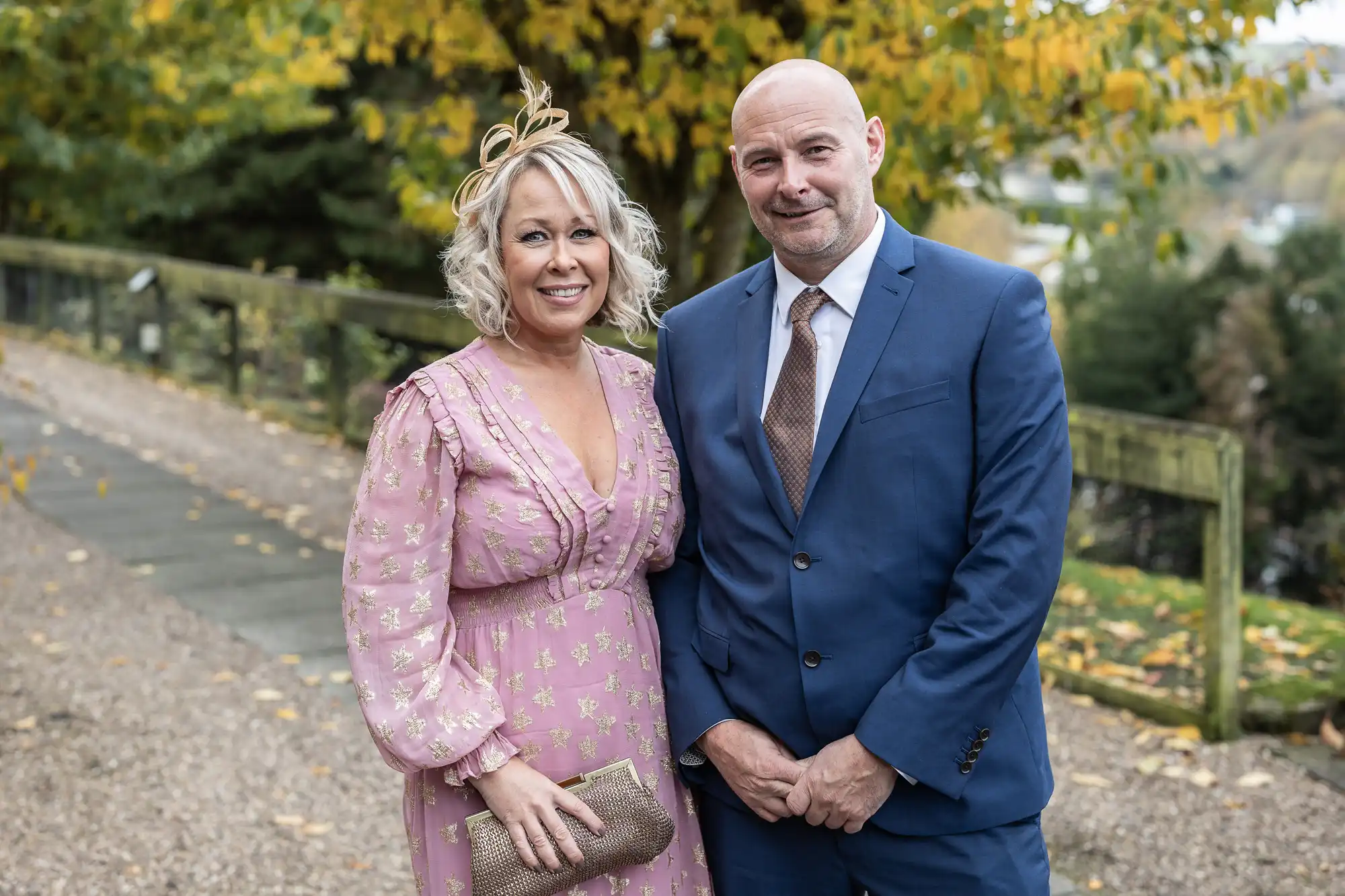 A smiling woman in a pink floral dress and a tan fascinator stands next to a bald man in a blue suit and brown tie. They pose outdoors on a gravel path with green trees and autumn leaves in the background.