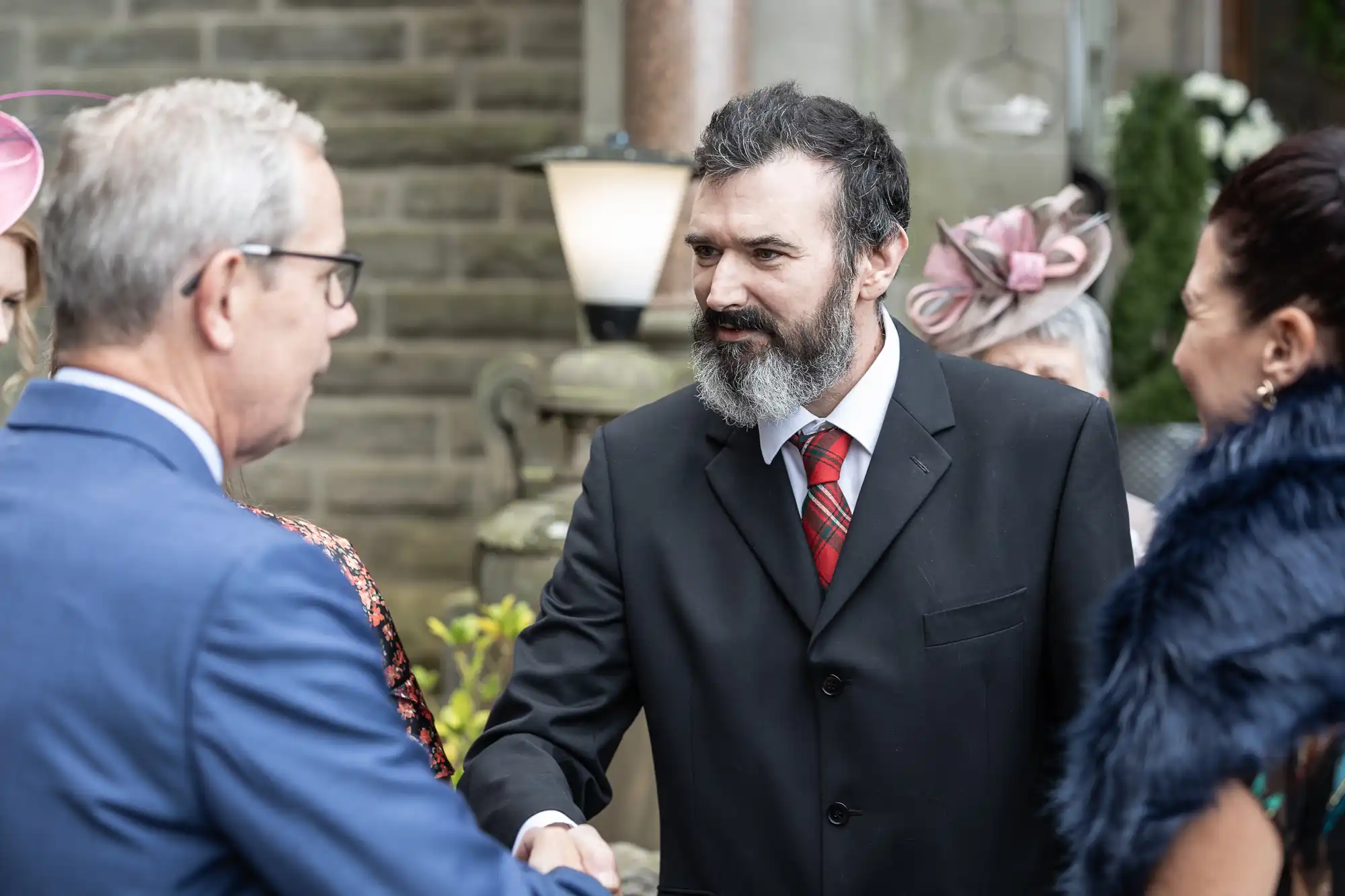 A bearded man in a black suit and red checked tie shakes hands with an older man in glasses and a blue suit at an outdoor formal event. Two women in elegant attire stand nearby. Stone walls and greenery are visible in the background.