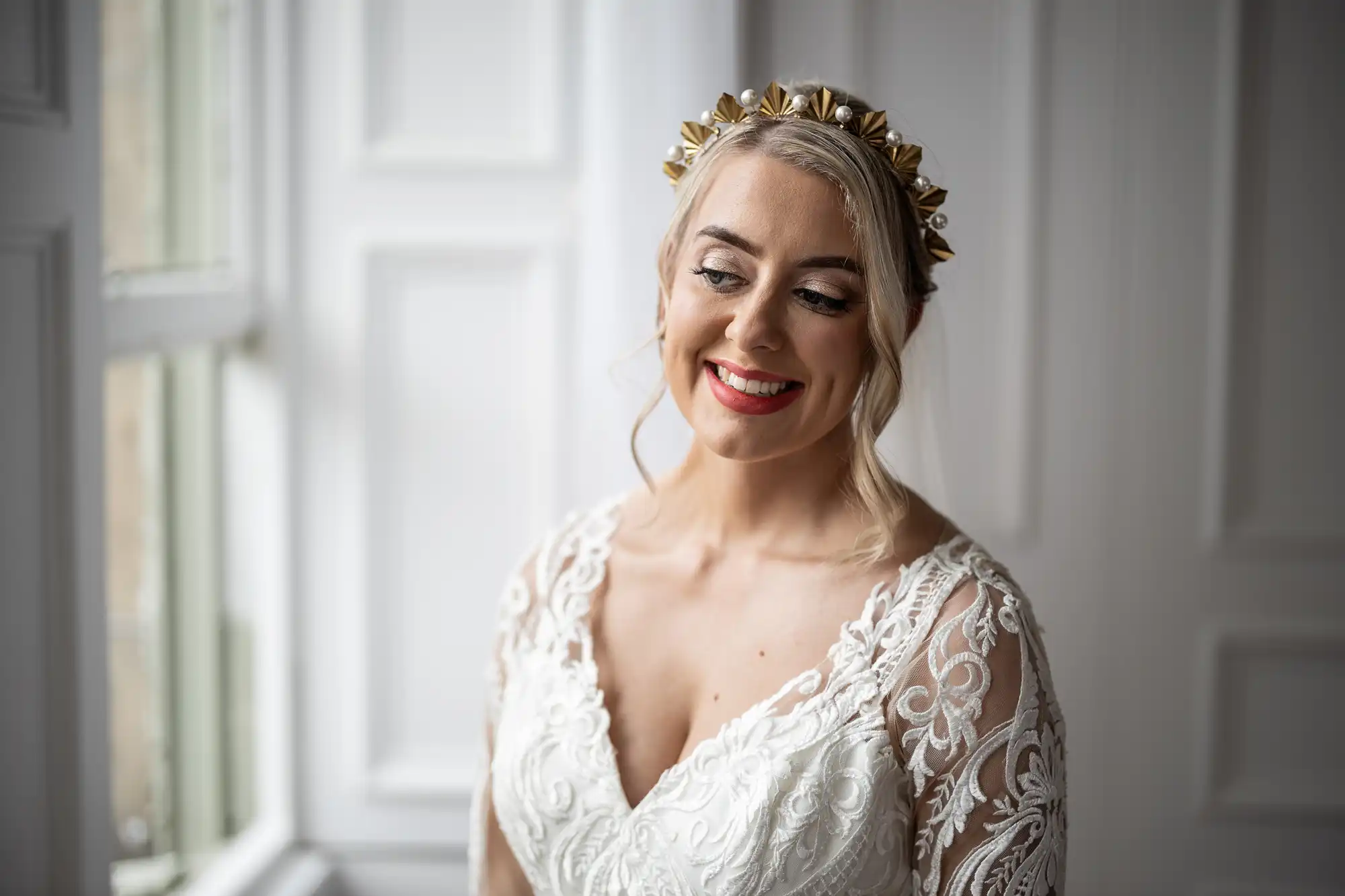 A smiling bride stands by a window in soft natural light, wearing a lace wedding dress with a deep V-neck and sheer embroidered sleeves. She has blonde hair styled up with loose strands and a gold spiked headpiece, and wears bright red lipstick.