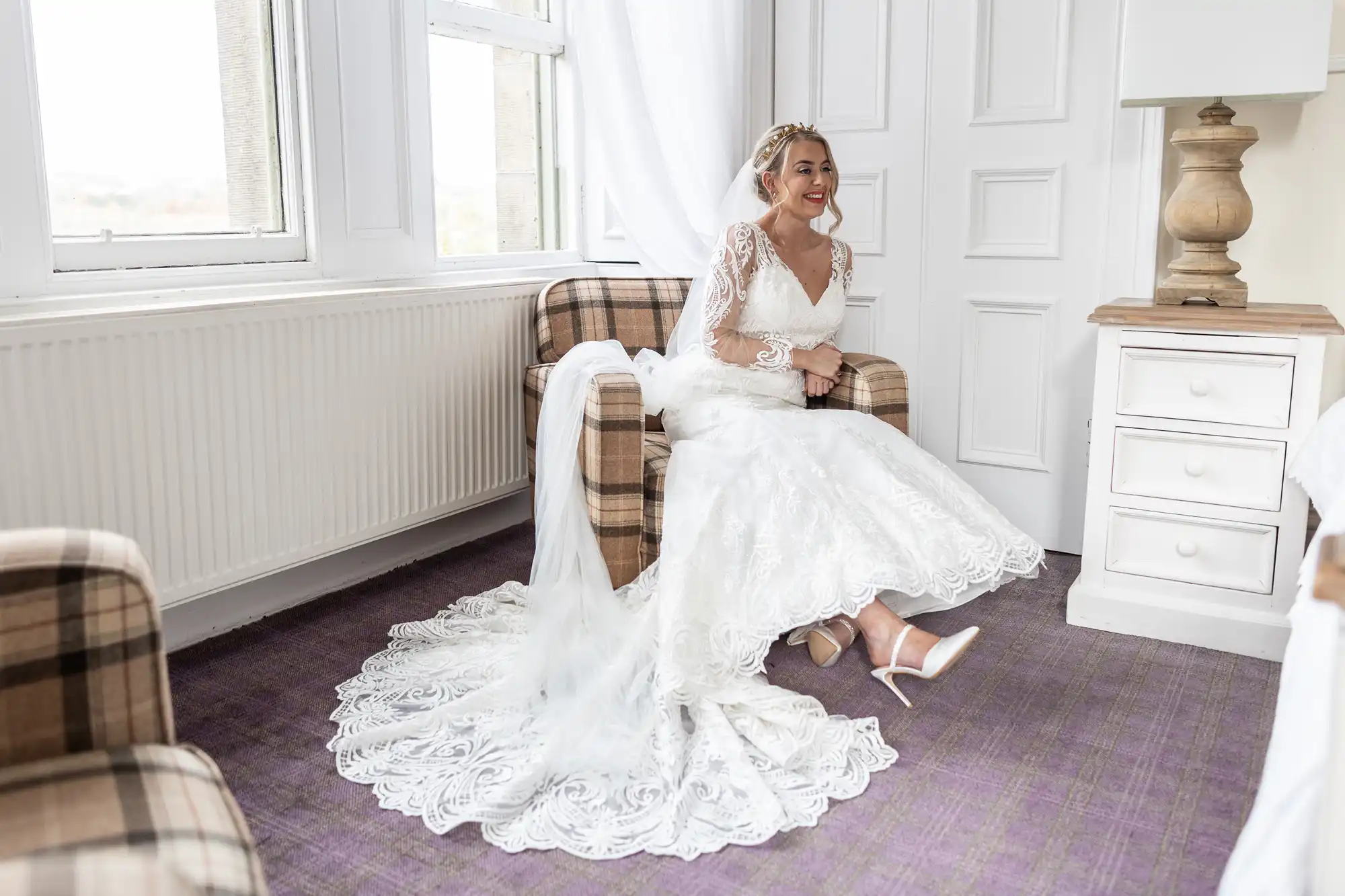 A smiling bride in a white lace wedding dress with a long train sits on a checked armchair in a bright, cosy room. She wears white heels and a veil, with her hair styled up. White walls, a window with sheer curtains, and a wooden bedside table are visible.