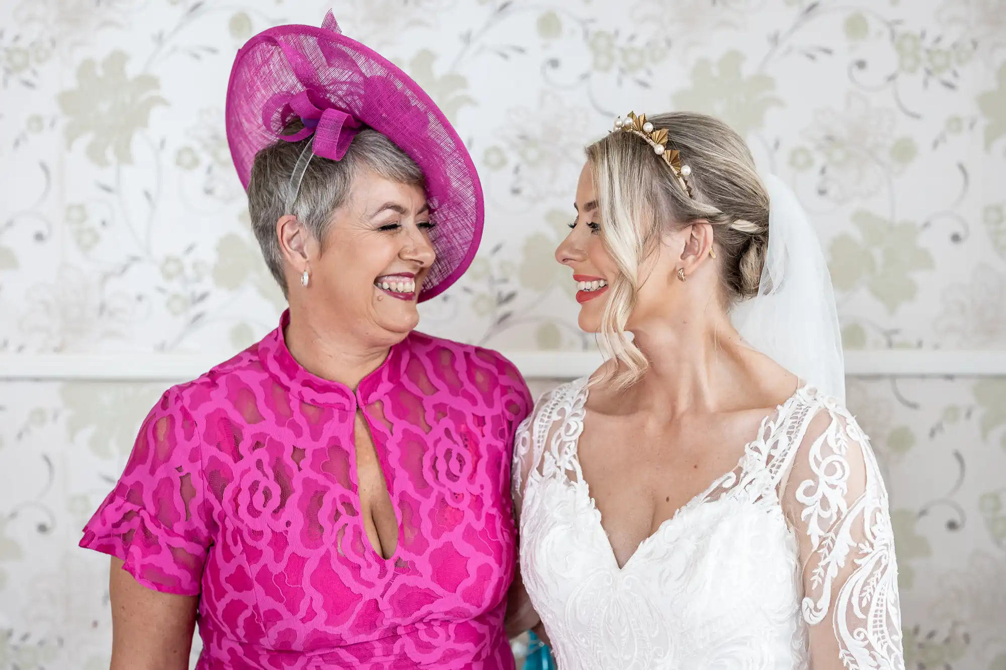 Two women smiling at each other. One wears a bright pink dress with a matching hat, and the other is in a white lace wedding dress with a veil and a gold headpiece. They stand against a light floral-patterned wall, appearing joyful and affectionate.