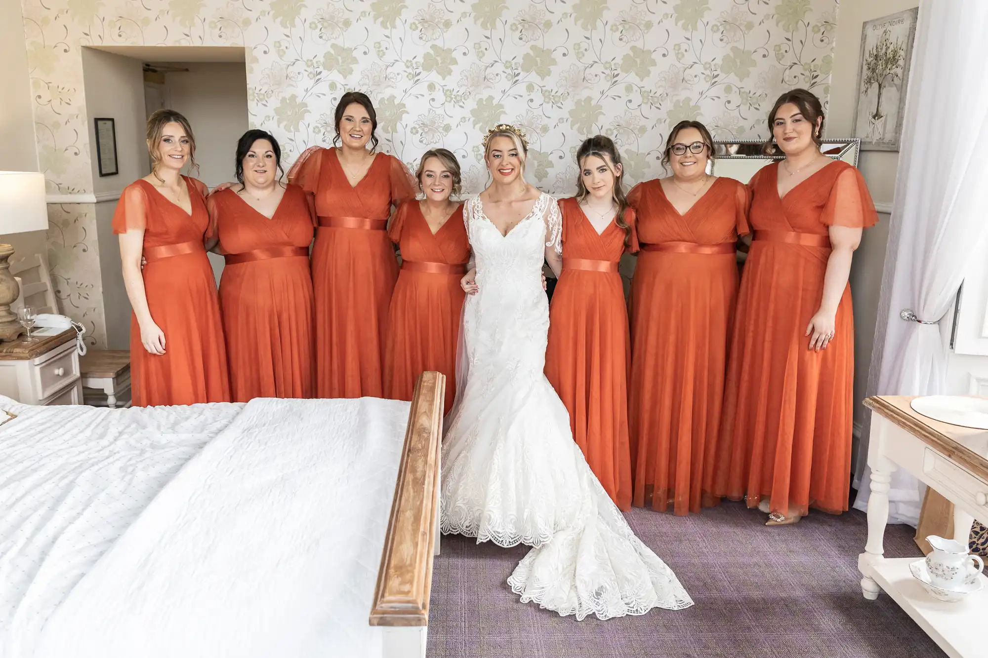 Eight women stand indoors. Seven bridesmaids wear matching burnt orange dresses, while the bride, at the centre, wears a white lace gown. They all smile, arms linked, in a softly lit bedroom with floral wallpaper and a made bed in the foreground.