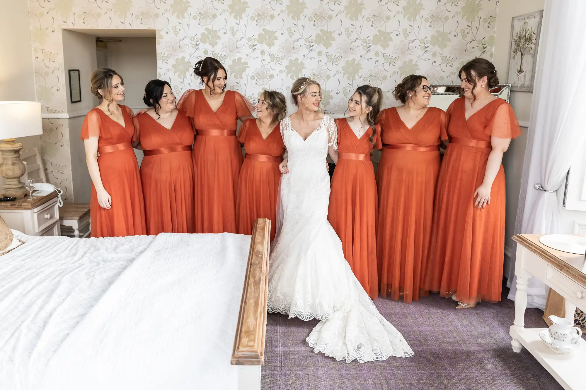 A bride in a white lace gown stands smiling and arm-in-arm with six bridesmaids in matching orange dresses. They stand indoors by a bed in a softly lit room with floral wallpaper, sharing a joyful moment together.