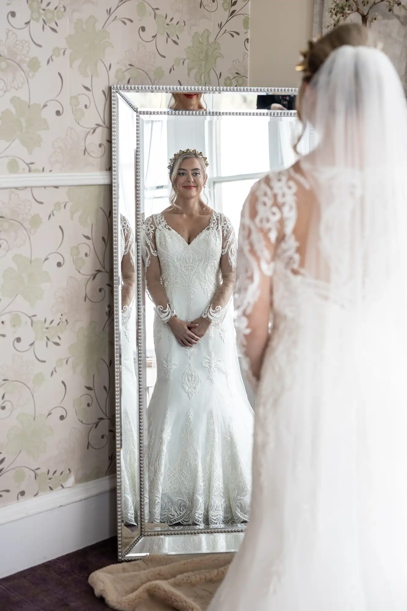 A bride in a white lace wedding dress and veil stands facing a tall mirror. Her reflection, showing her smiling and holding her hands together, is visible in the mirror. The room has floral wallpaper and soft light from the side.