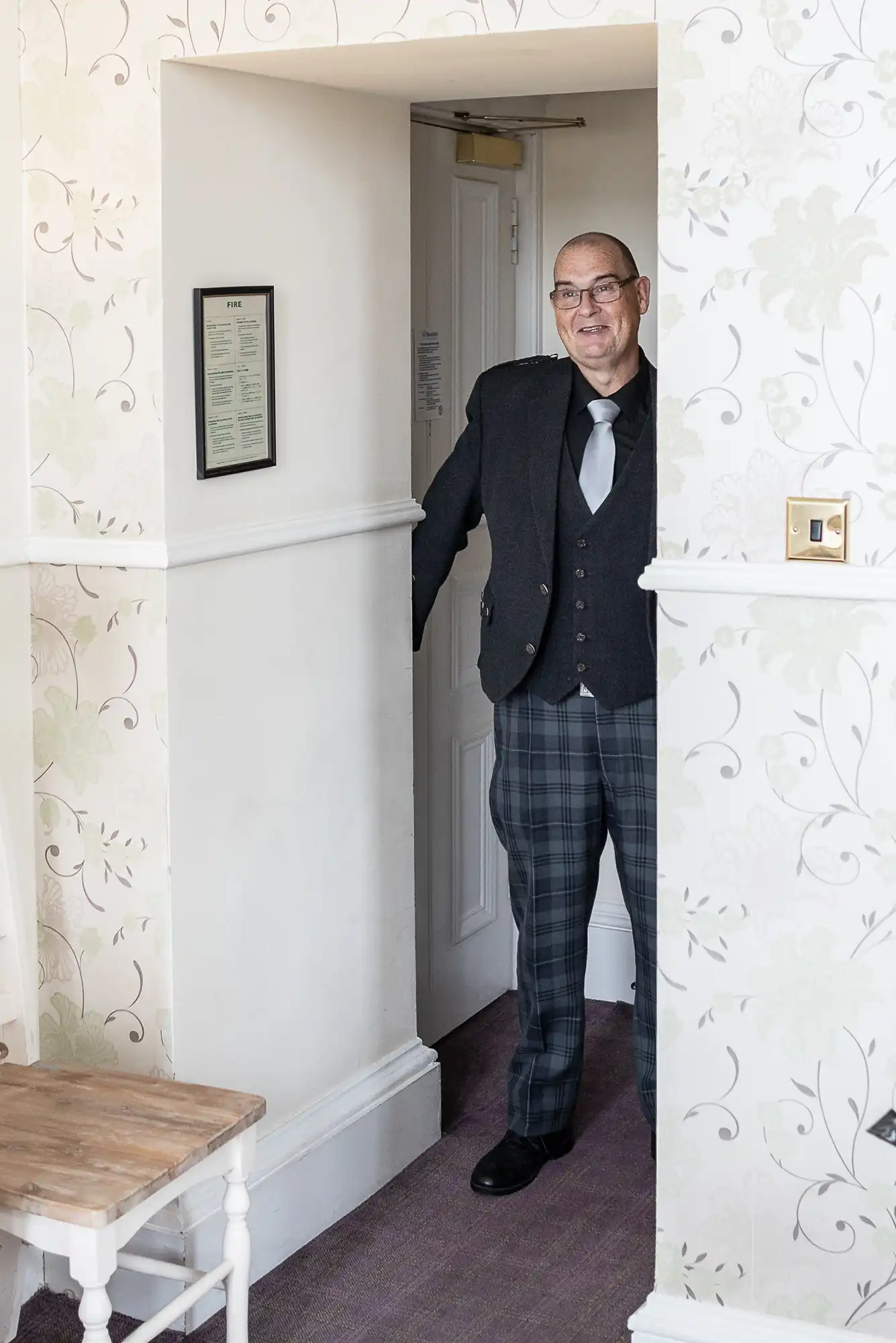 A smiling man with glasses, wearing a dark jacket, grey tie, and checked trousers, stands in a doorway. The room has light floral wallpaper, a wooden bench, and a framed notice on the wall. The carpet is purple, and light fills the space.