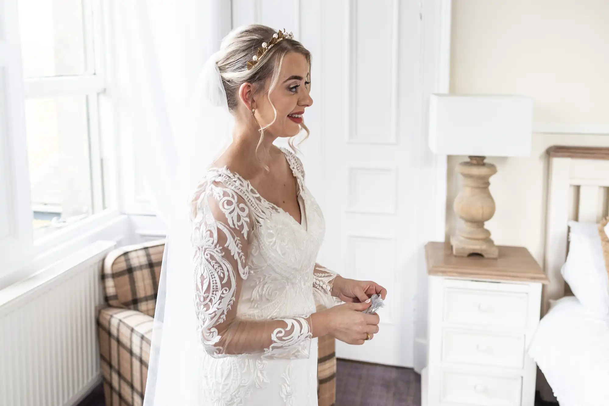 A bride in a white lace wedding dress with sheer long sleeves stands in a bright room, smiling. She has a veil and pearl headdress. The room features white walls, a lamp on a wooden bedside table, a bed, and a checked armchair.