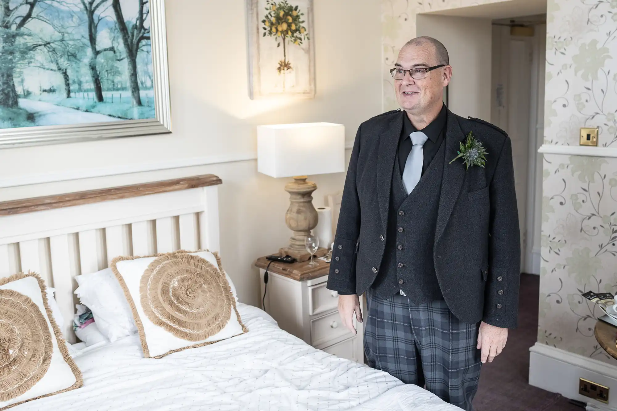 A man with glasses, wearing a dark jacket, waistcoat, plaid kilt, and tie with a buttonhole, stands smiling beside a bed in a softly lit, elegantly decorated bedroom with light walls, art, and patterned cushions.