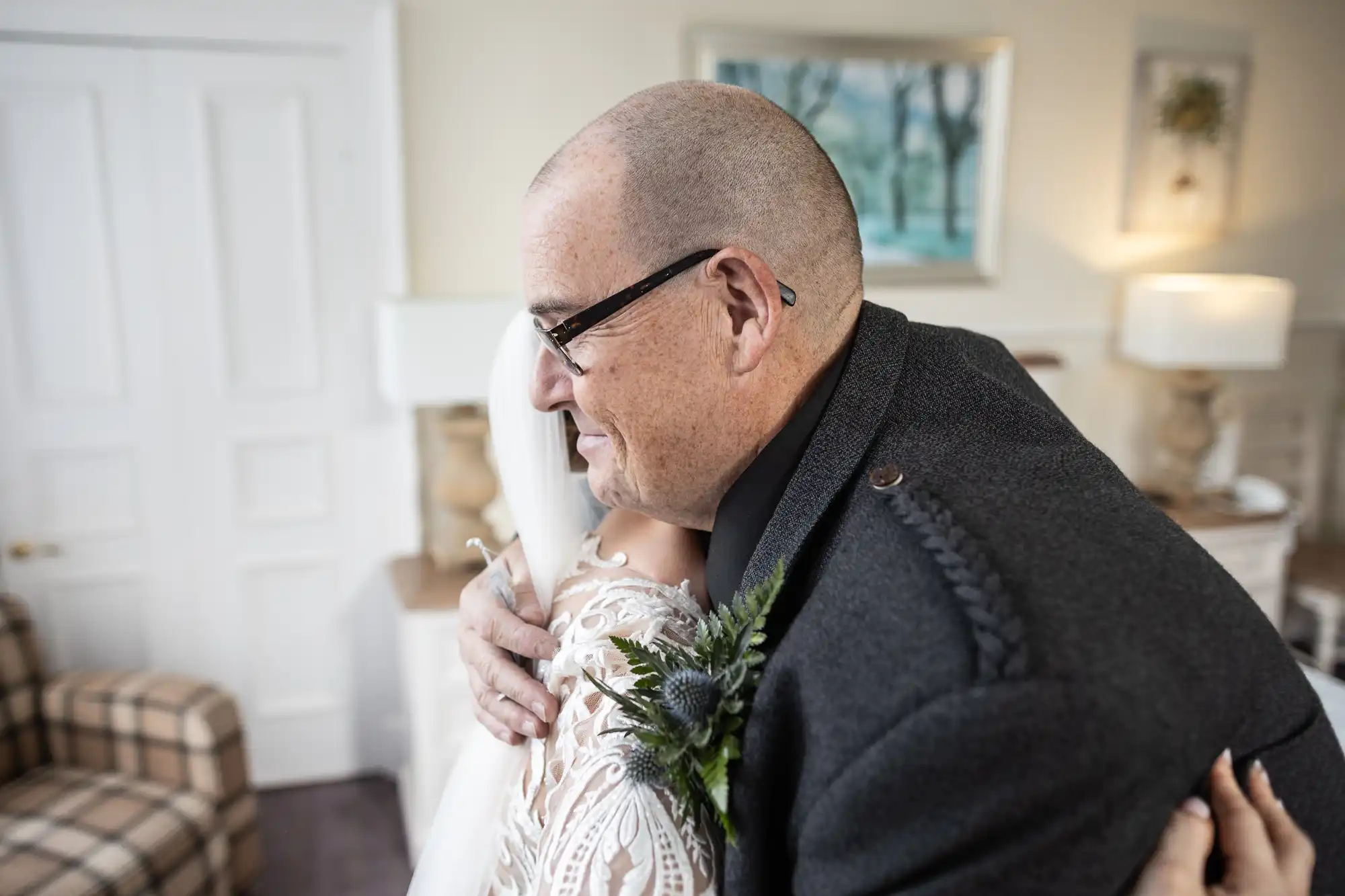 A man with glasses and a shaven head hugs a woman in a white lace dress. The scene appears warm and emotional, set in a bright room with paintings, lamps, and a checked armchair in the background. The woman holds a tissue in her hand.