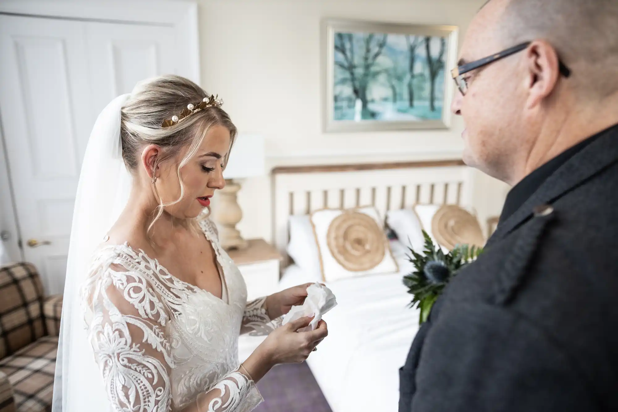 A bride in a lace wedding dress and veil stands indoors, holding a handkerchief and looking down emotionally. A man in glasses, likely her father, stands nearby. The scene takes place in a softly lit bedroom with a made bed and framed art on the wall.