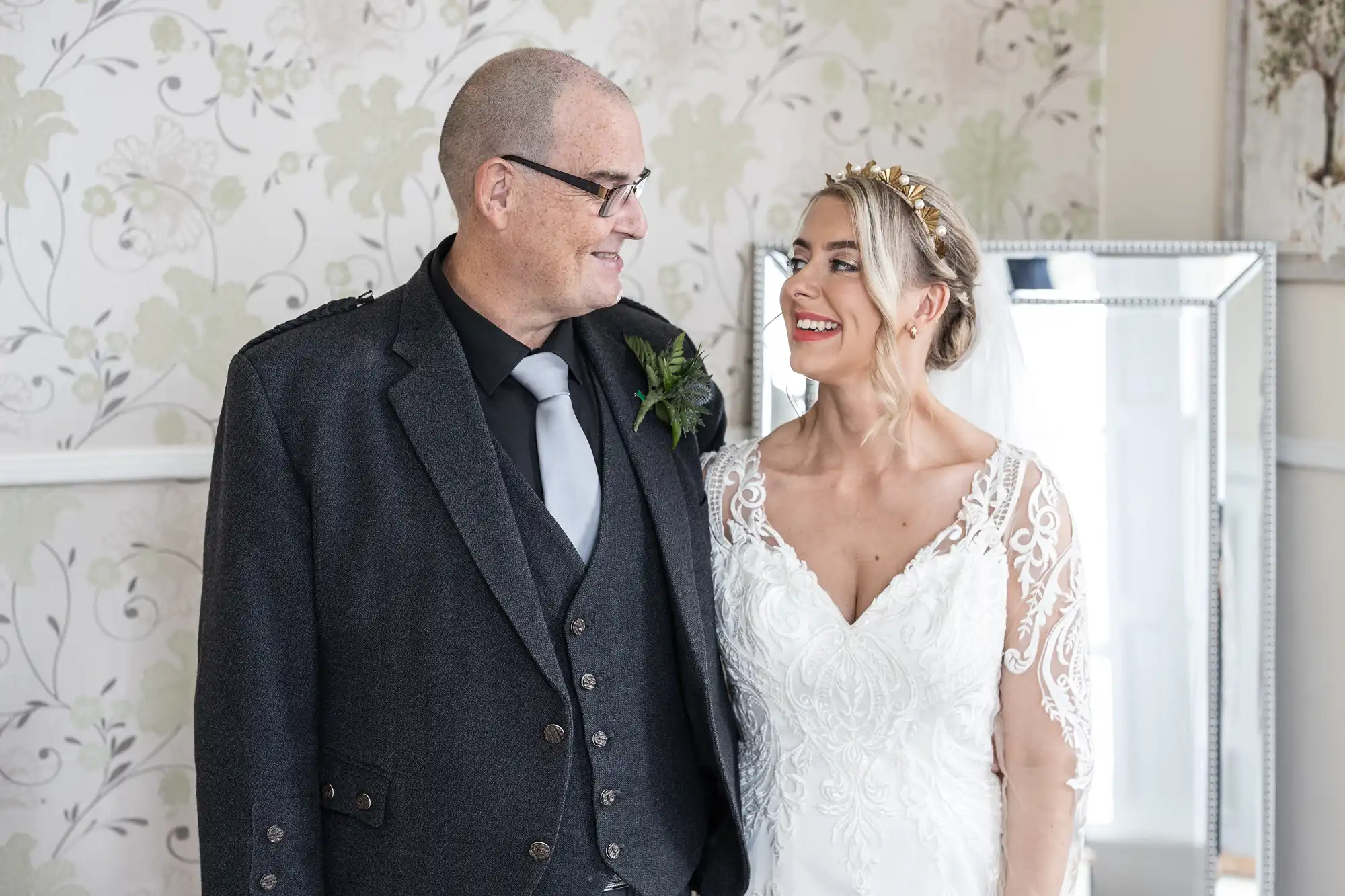 A smiling bride in a white lace dress and a tiara stands next to an older man in glasses, a dark suit, and a grey tie. They face each other, smiling warmly, in a bright room with floral wallpaper and a large mirror in the background.
