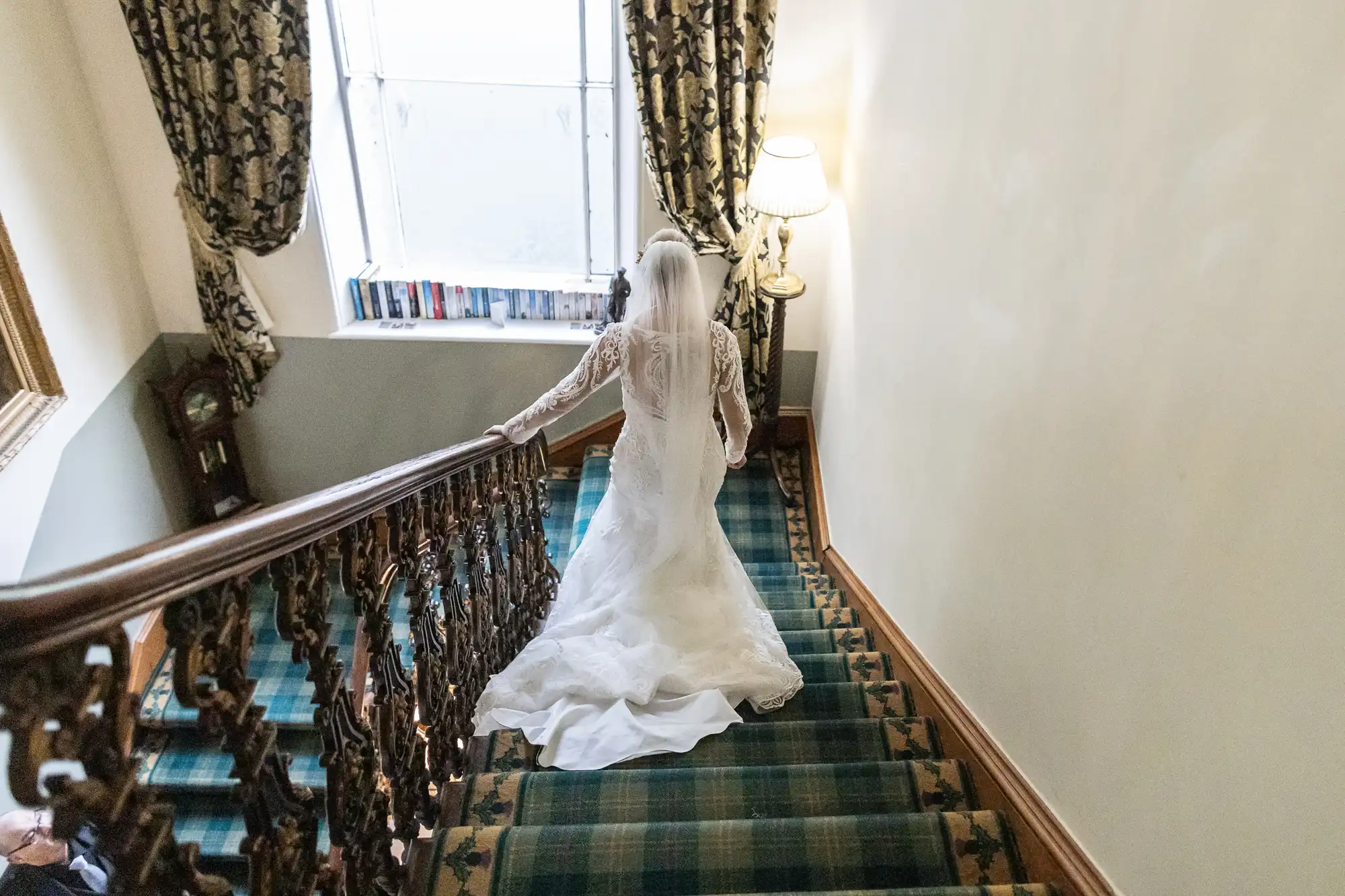 A bride in a white wedding dress and veil descends a carpeted staircase with ornate banister. Sunlight streams through a window, illuminating the scene. Patterned curtains and a standard lamp decorate the elegant interior.