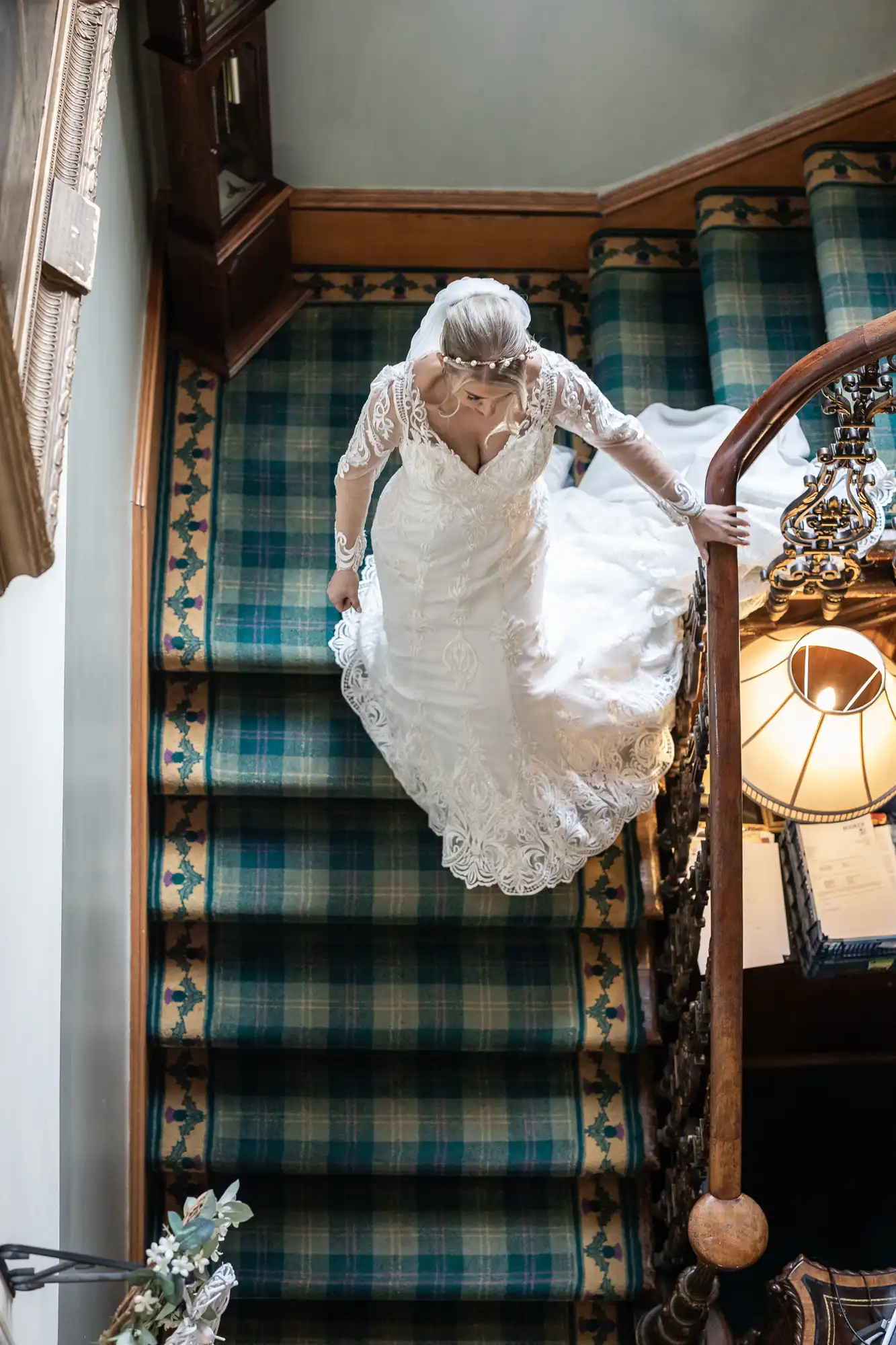 A bride in a white lace gown with long sleeves descends a staircase covered in green and blue tartan carpet. She holds the bannister and looks down, her veil trailing behind her. A decorative lamp and framed art are visible nearby.
