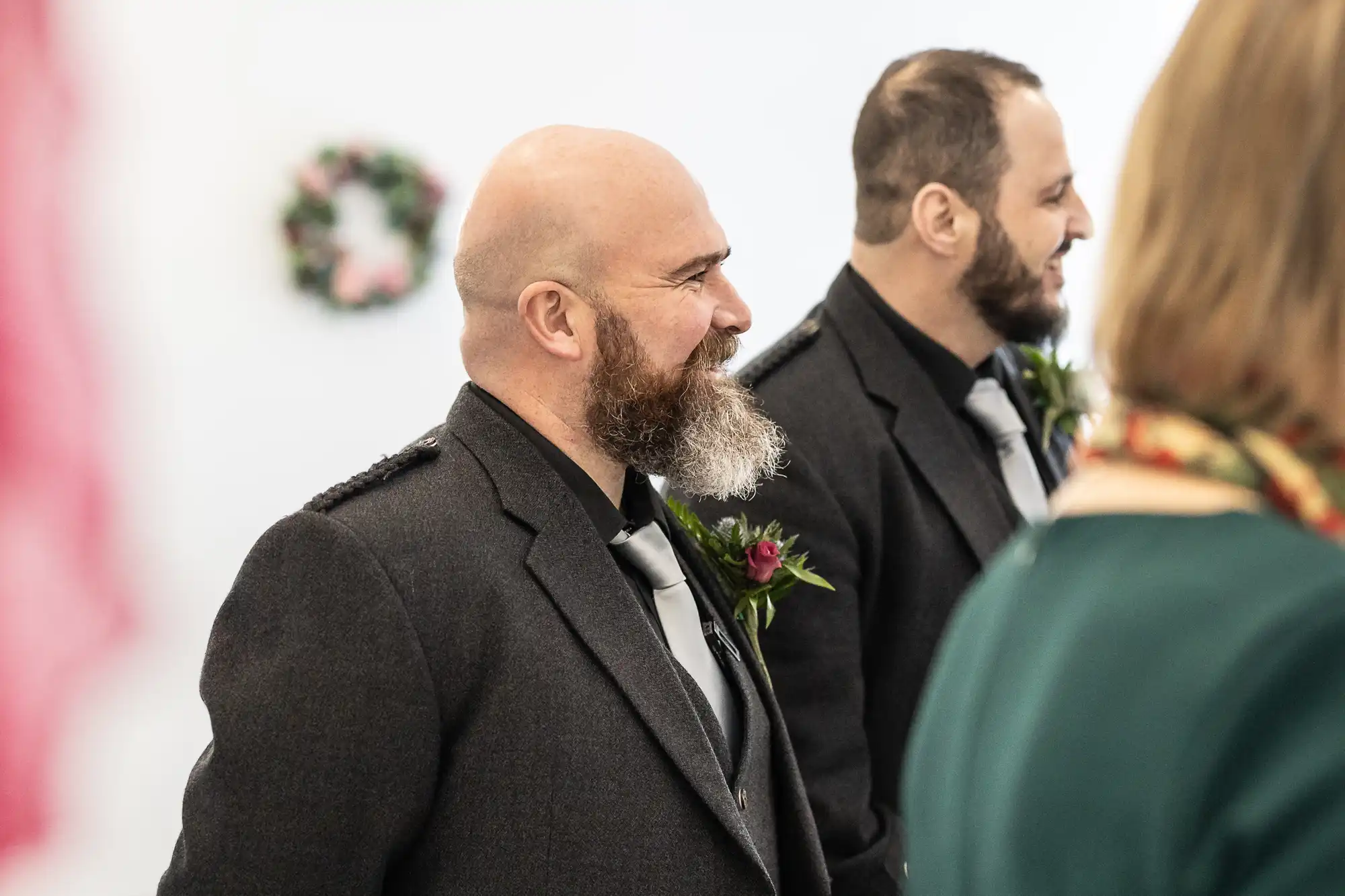 Two men in dark suits with buttonholes and matching silver ties stand indoors, smiling. Both have beards and short hair. A blurred person in green and a floral wreath on a white wall are visible in the background.